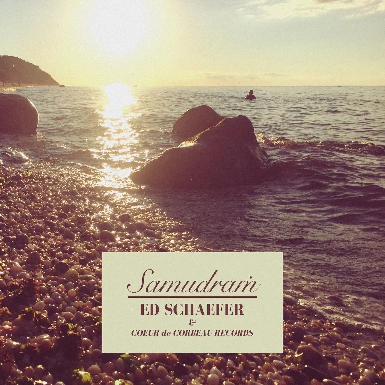 A beach scene at sunset with rocks in the foreground, a person swimming in the distance, and the sun reflecting off the water.