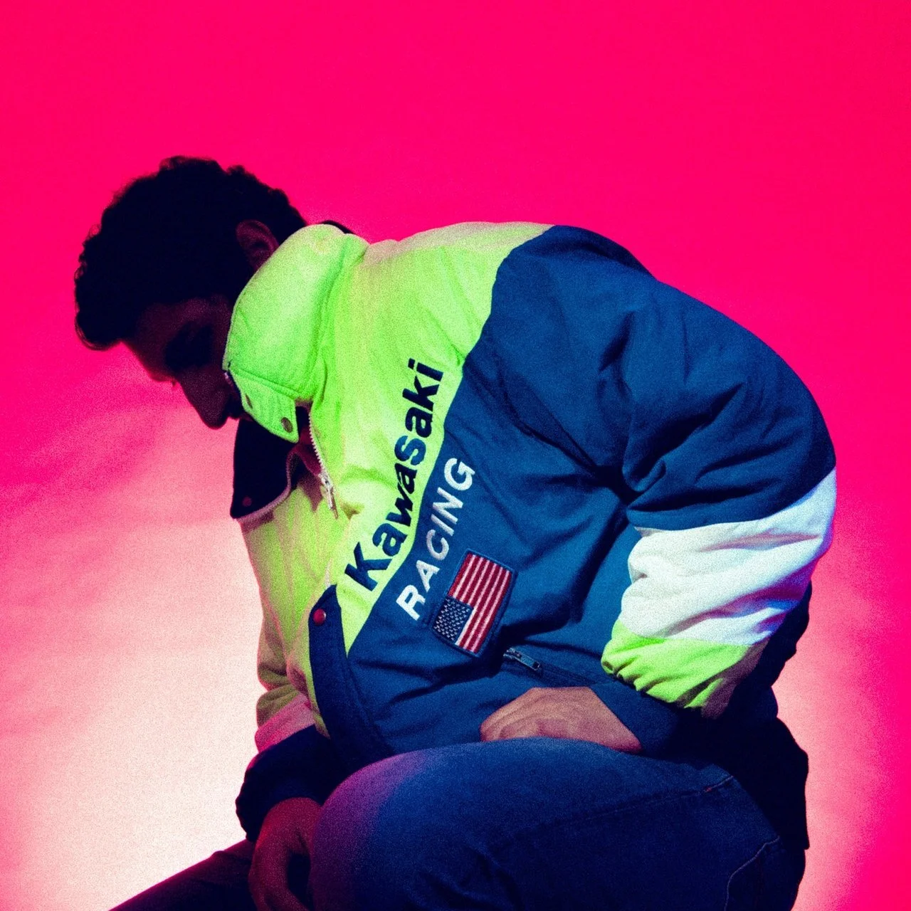 Young man crouching against a pink background, wearing a neon yellow and blue Kawasaki Racing jacket.