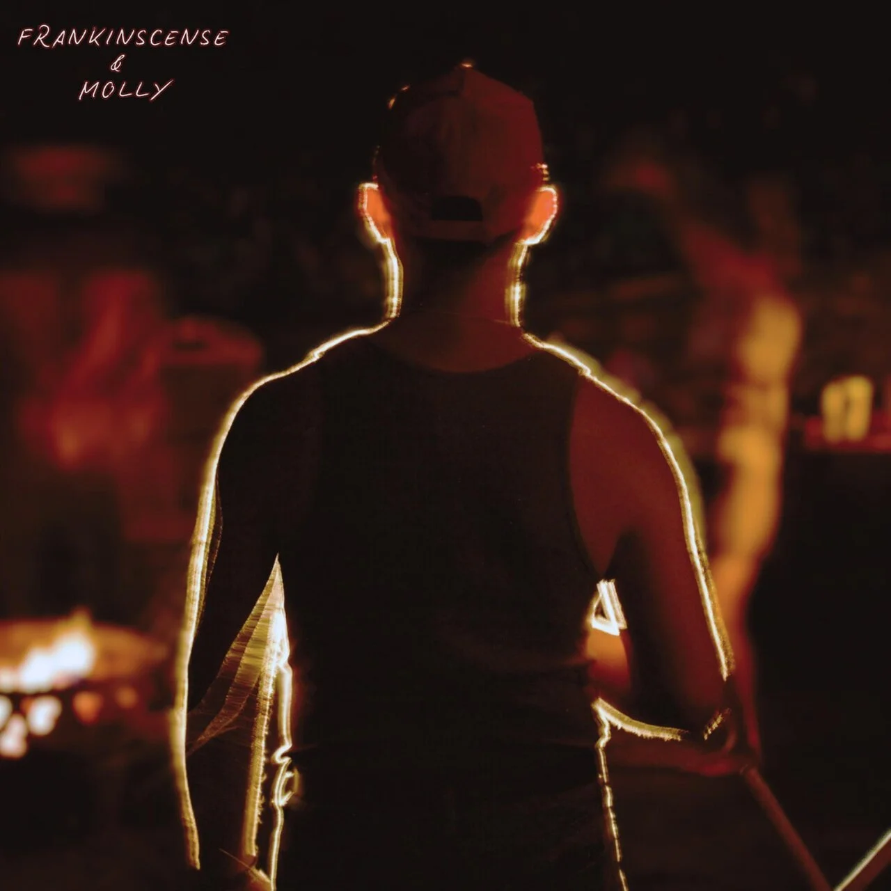 Person wearing a cap and sleeveless shirt, illuminated by red and yellow lights, standing outdoors at night with others in the background.