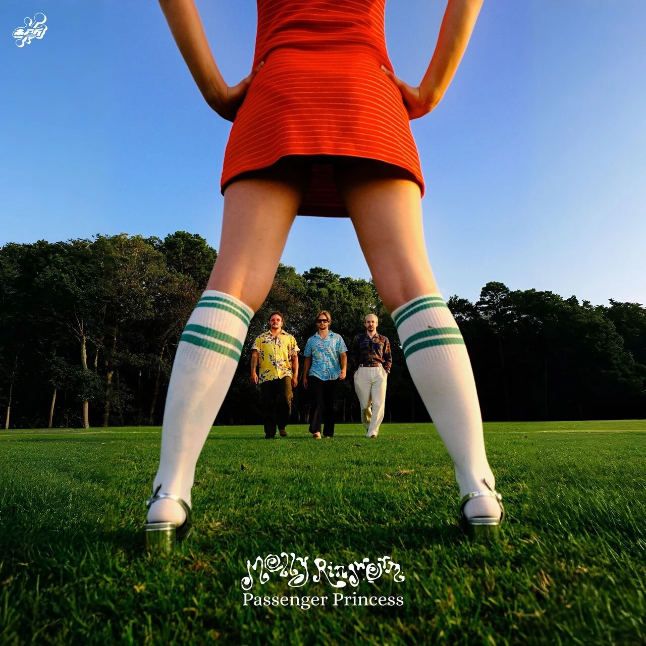 A person in an orange dress and knee-high socks stands with hands on hips in the foreground, looking at three men walking towards them on a grassy field with trees in the background during sunset.