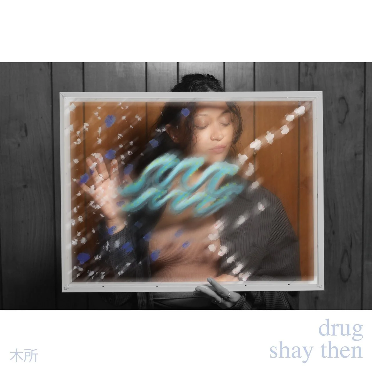 A woman with dark, curly hair holding a glass frame in front of her face. Inside the frame, graffiti-like colorful text appears, while she is surrounded by black, wooden paneling.