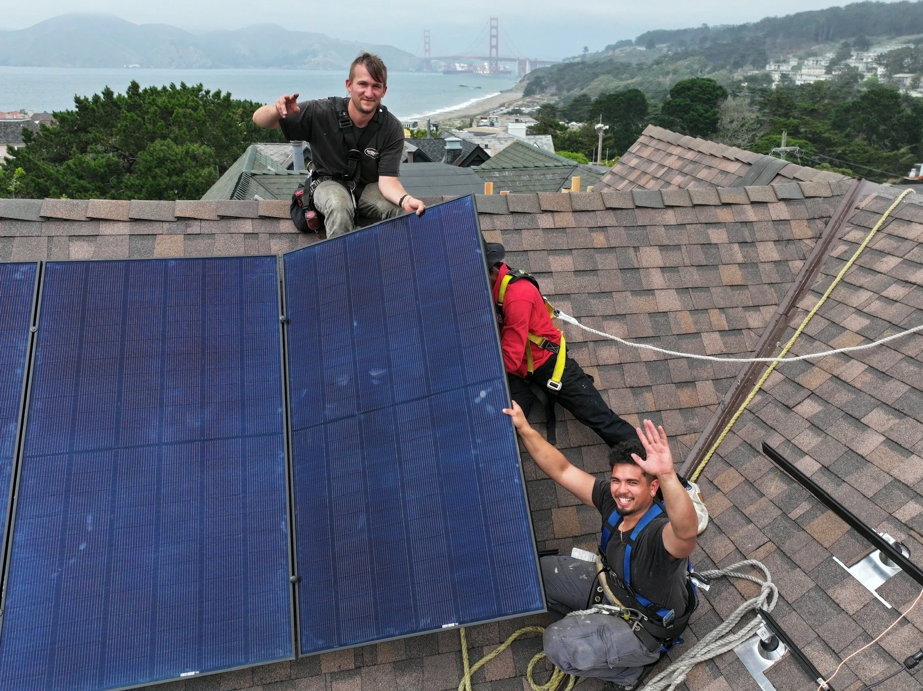 Two men installing solar panels on a house roof with a coastal landscape and the Golden Gate Bridge in the background.