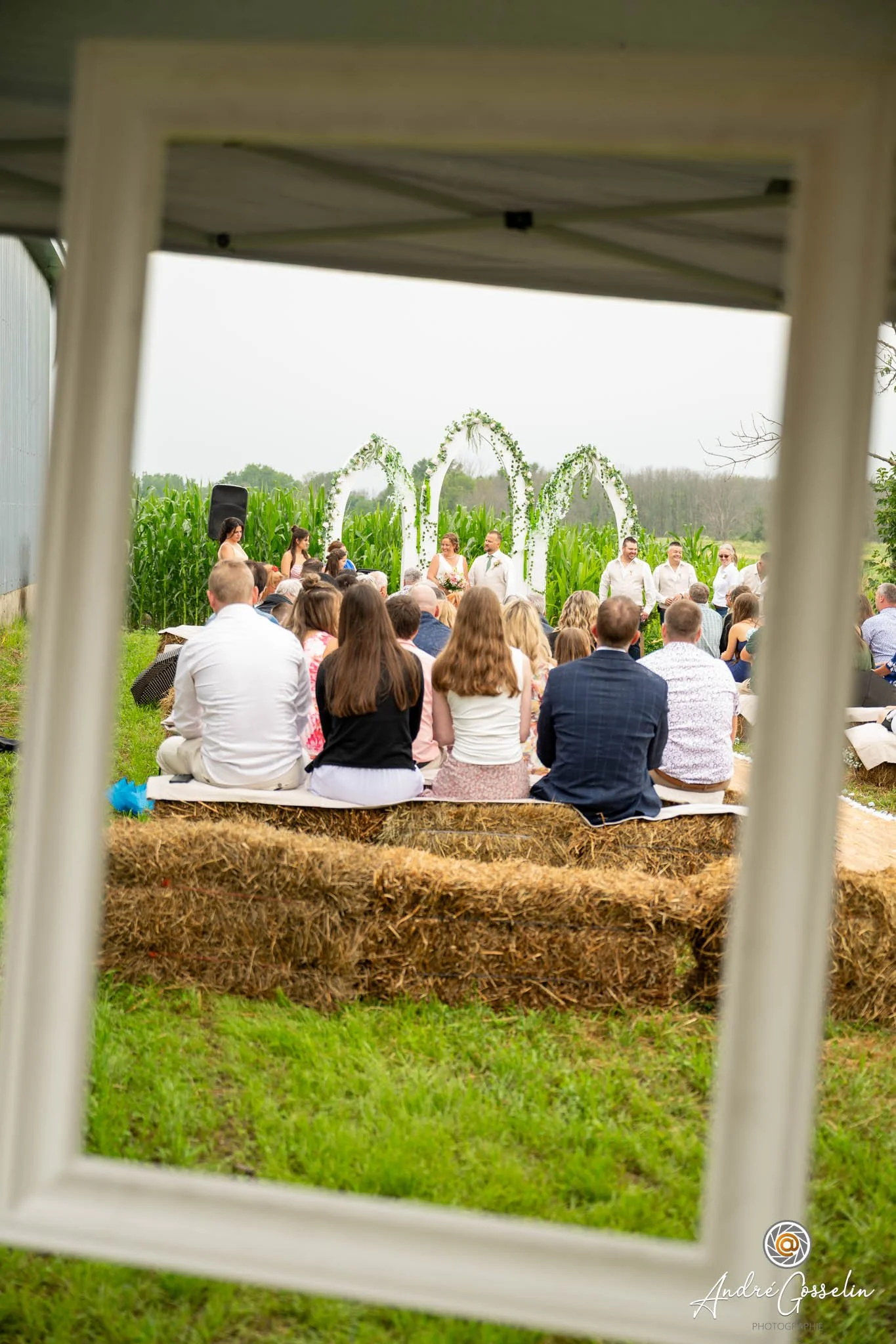 Cérémonie de mariage en plein air dans un champ de maïs avec des invités assis sur des bottes de foin, un couple marié et des officiants sous un arc décoré de fleurs.