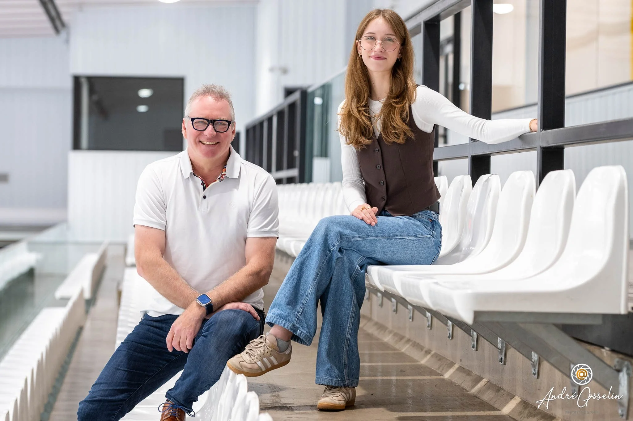 Deux personnes assises sur des bancs blancs dans une salle intérieure. Une homme âgé souriant portant un polo blanc et un homme plus jeune avec un sweat à manches longues blanc et une veste marron.