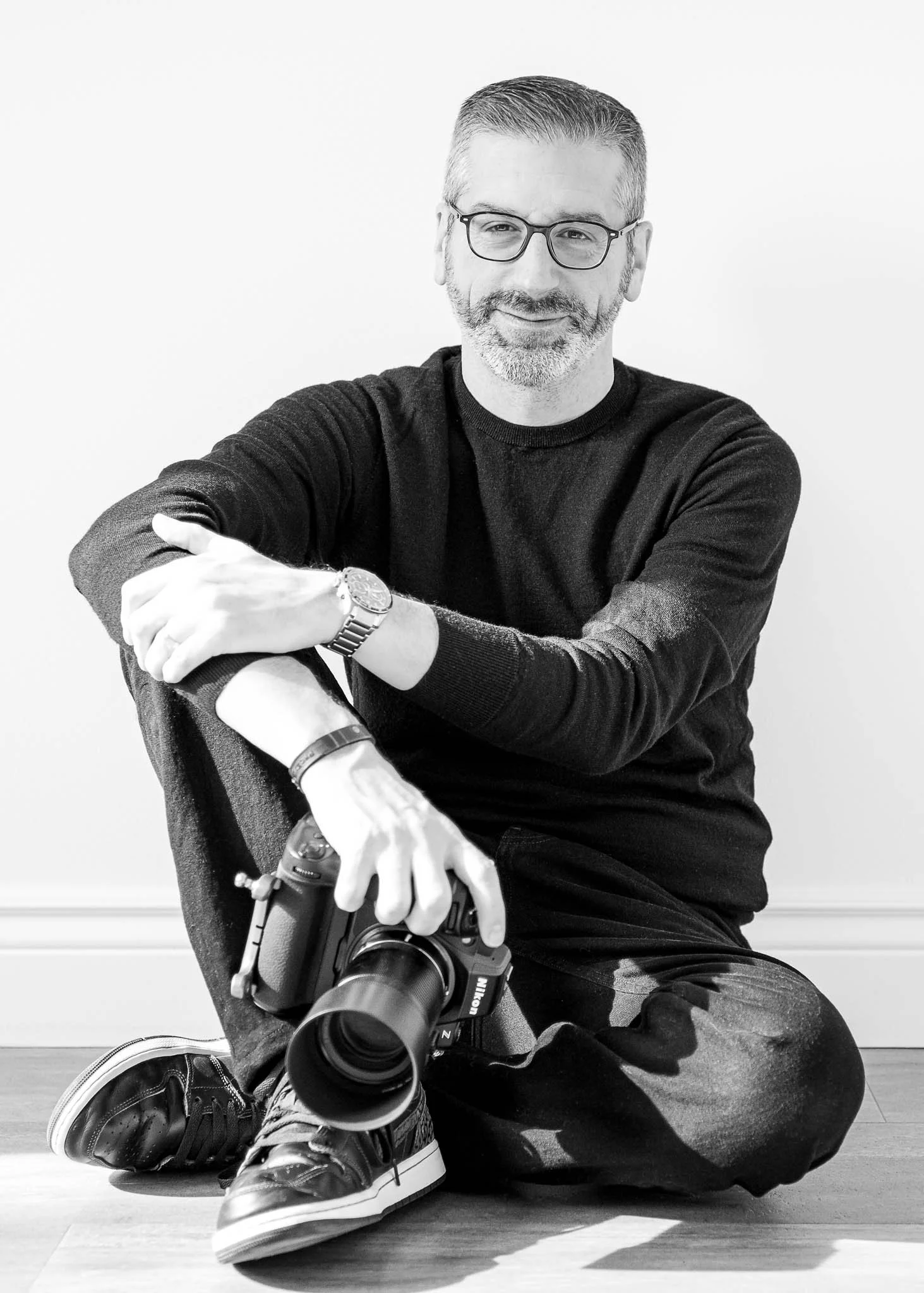 André Gosselin. Photographe de portrait. Photographe événement. Photo corporative. Homme assis par terre, tenant un appareil photo, portant des lunettes, un pull noir, des sneakers noirs, un montre et un bracelet, sourire en regardant l'objectif