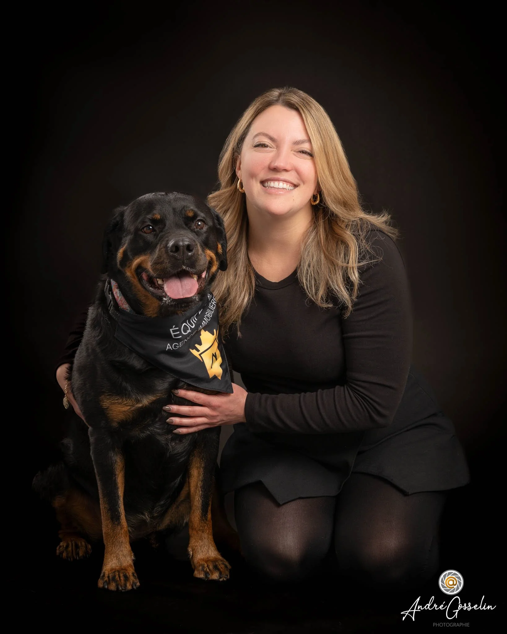 Une femme et un chien posent ensemble devant un fond noir. La femme sourit et porte des vêtements noirs, tandis que le chien, un Rottweiler, porte un bandana noir avec un logo et du texte en français.