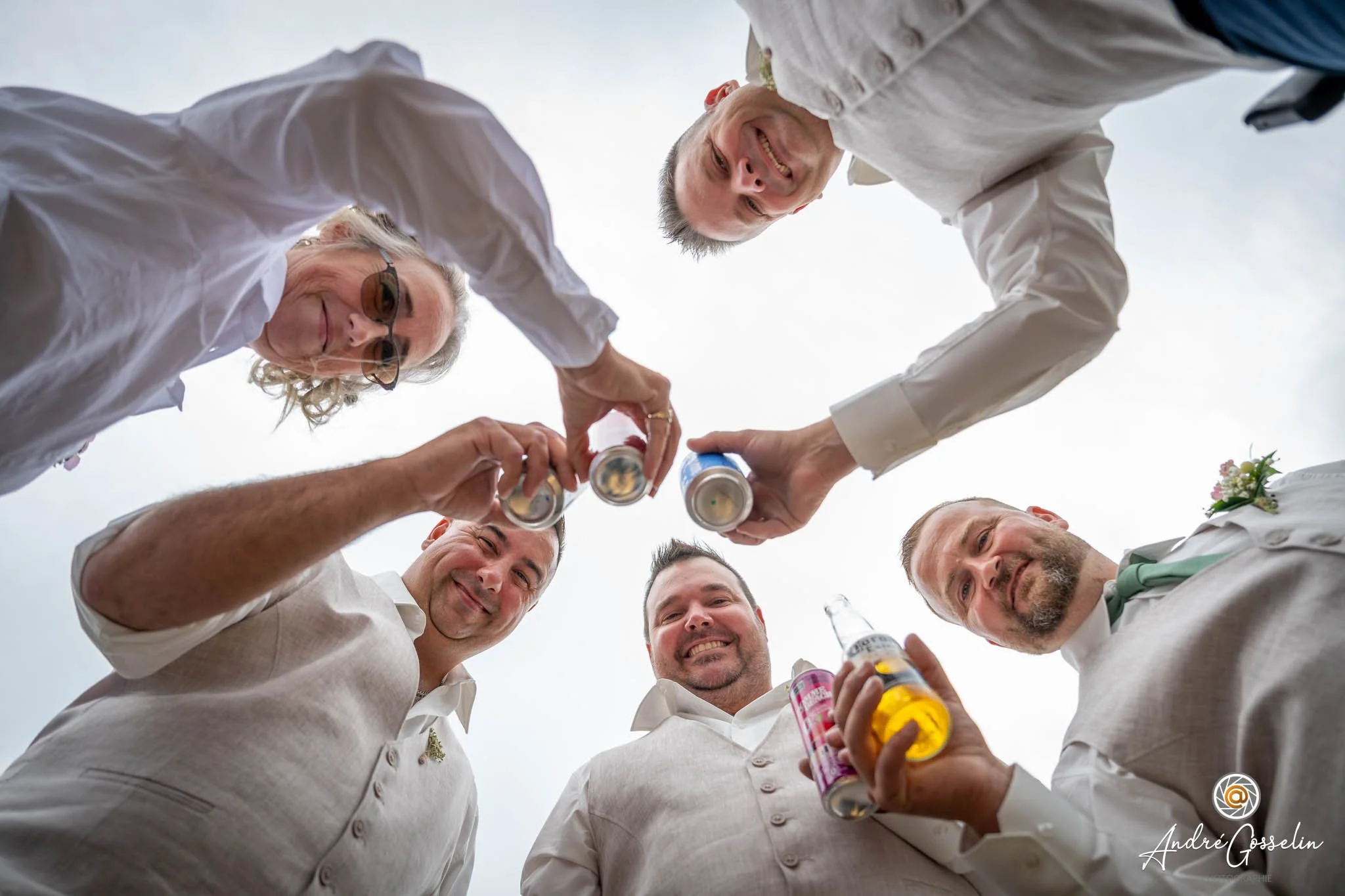 Groupe de six personnes souriantes lors d'une célébration, levant des canettes de boisson, photographiées en contre-plongée contre un ciel clair.