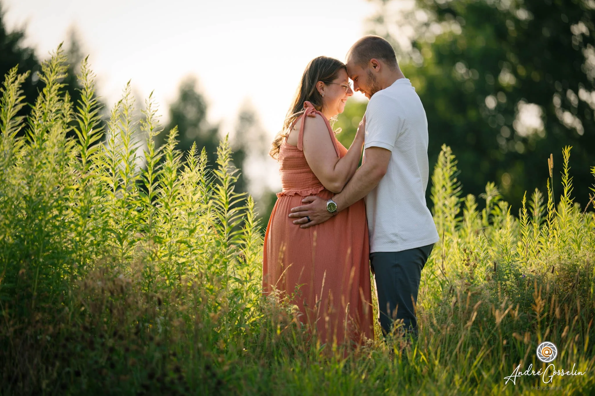 Un couple se tenant la tête-touchant leur front, souriant et regardant dans les yeux dans un champ avec des arbres en arrière-plan, au coucher de soleil. Séance de maternité au soleil couchant.