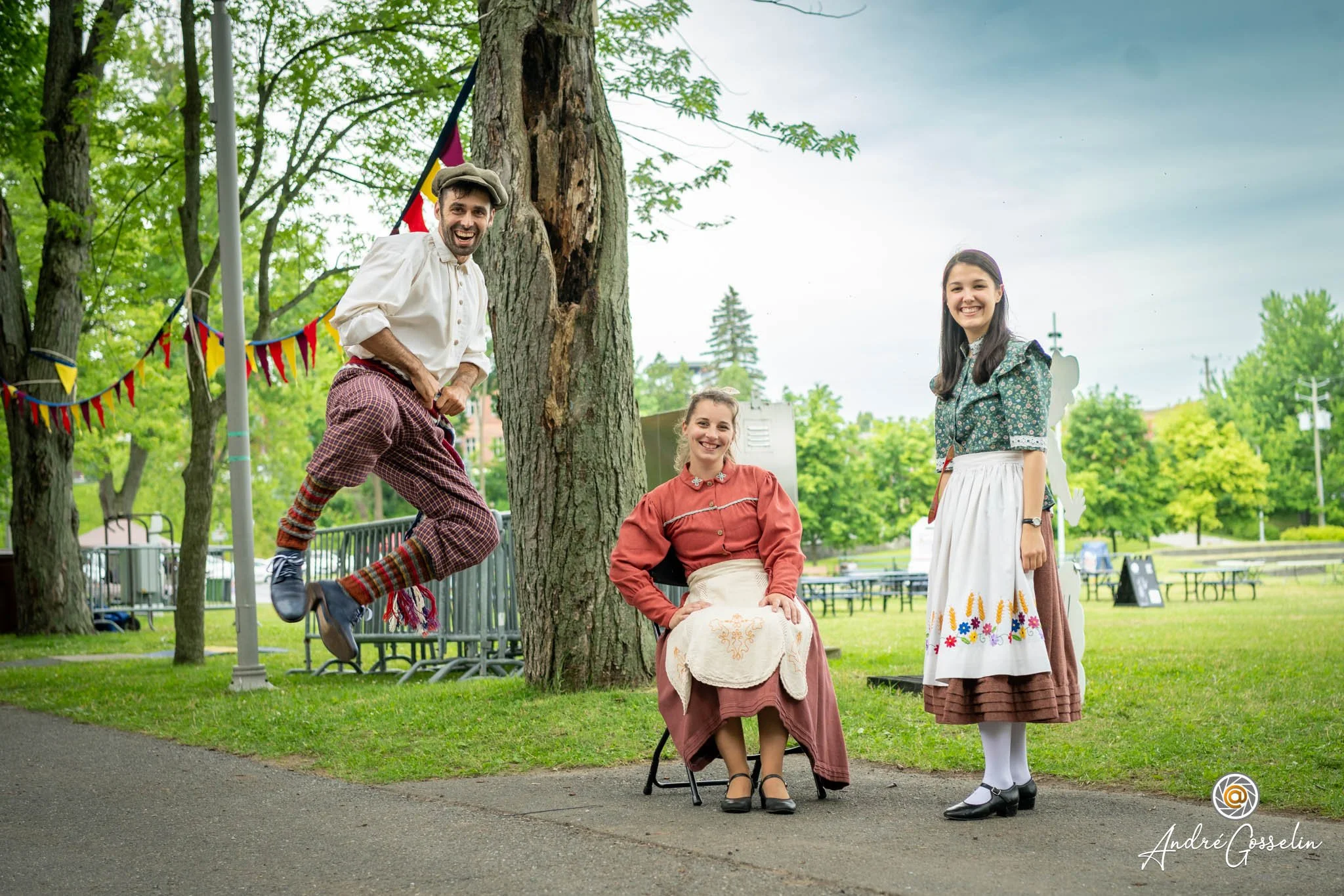 Trois personnes en costume traditionnel folklorique dans un parc, avec arbre et décorations colorées en arrière-plan, souriantes et posant pour la photo. Photo d'événement.