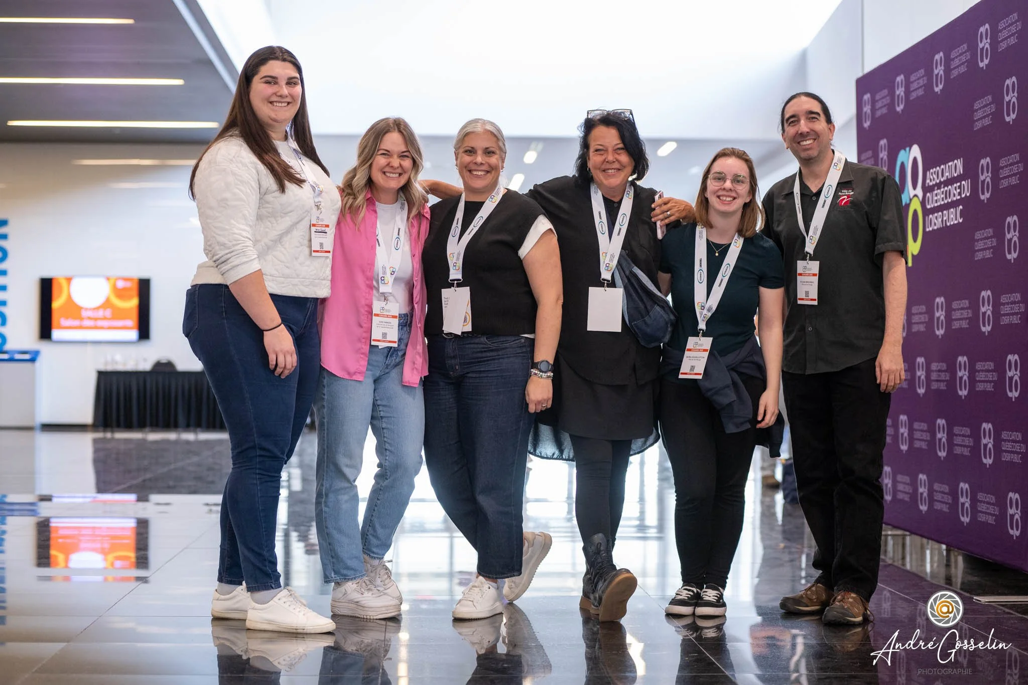 Groupe de six personnes souriantes en convention, portant des badges, devant un mur violet avec le logo de l'Association Québécoise du Loisir Public.