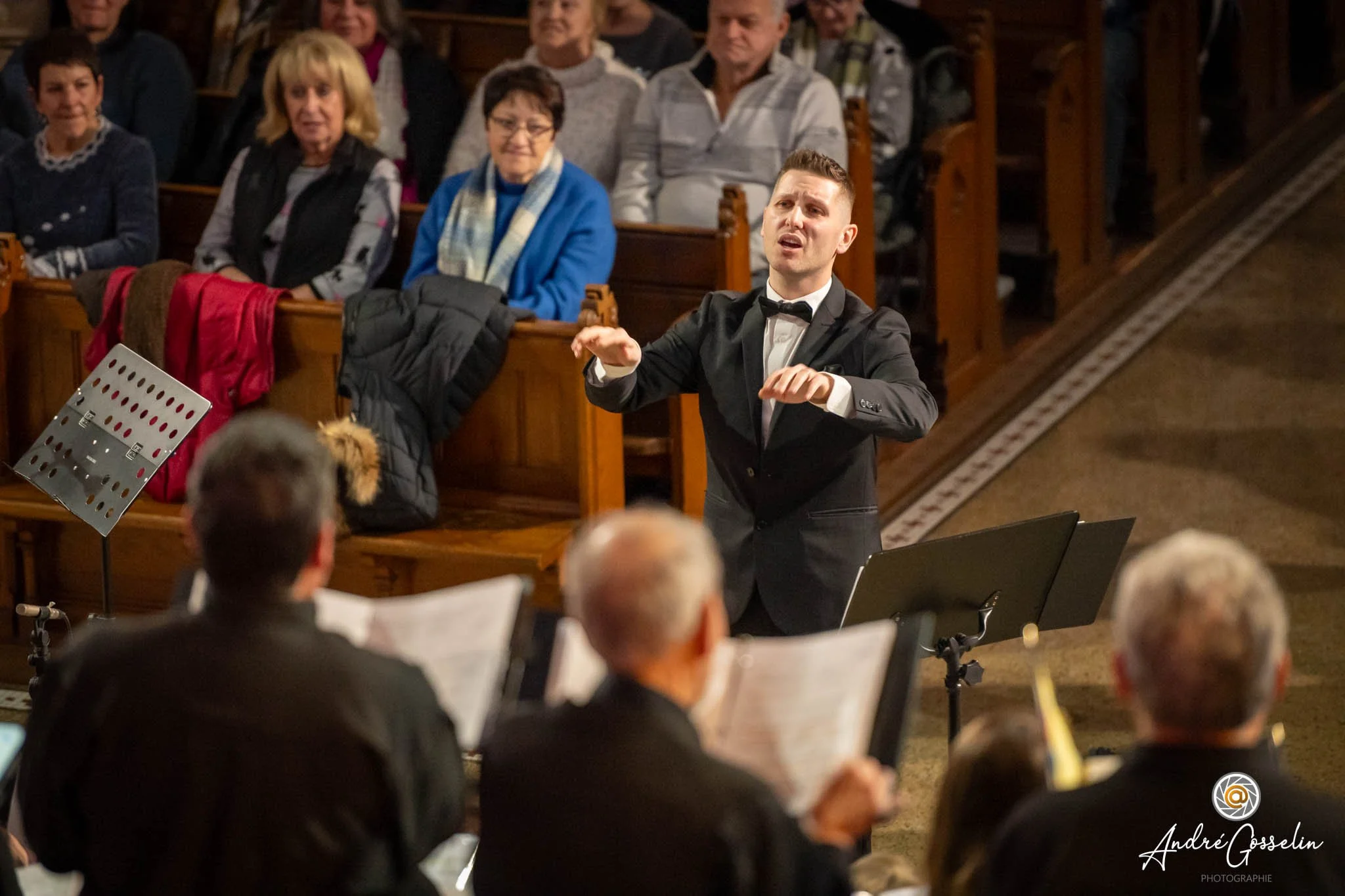 Un chef d'orchestre dirige un groupe de musiciens dans une salle de concert en bois, avec un public d'adultes assis dans des bancs en arrière-plan, certains regardant attentivement, d'autres souriant.