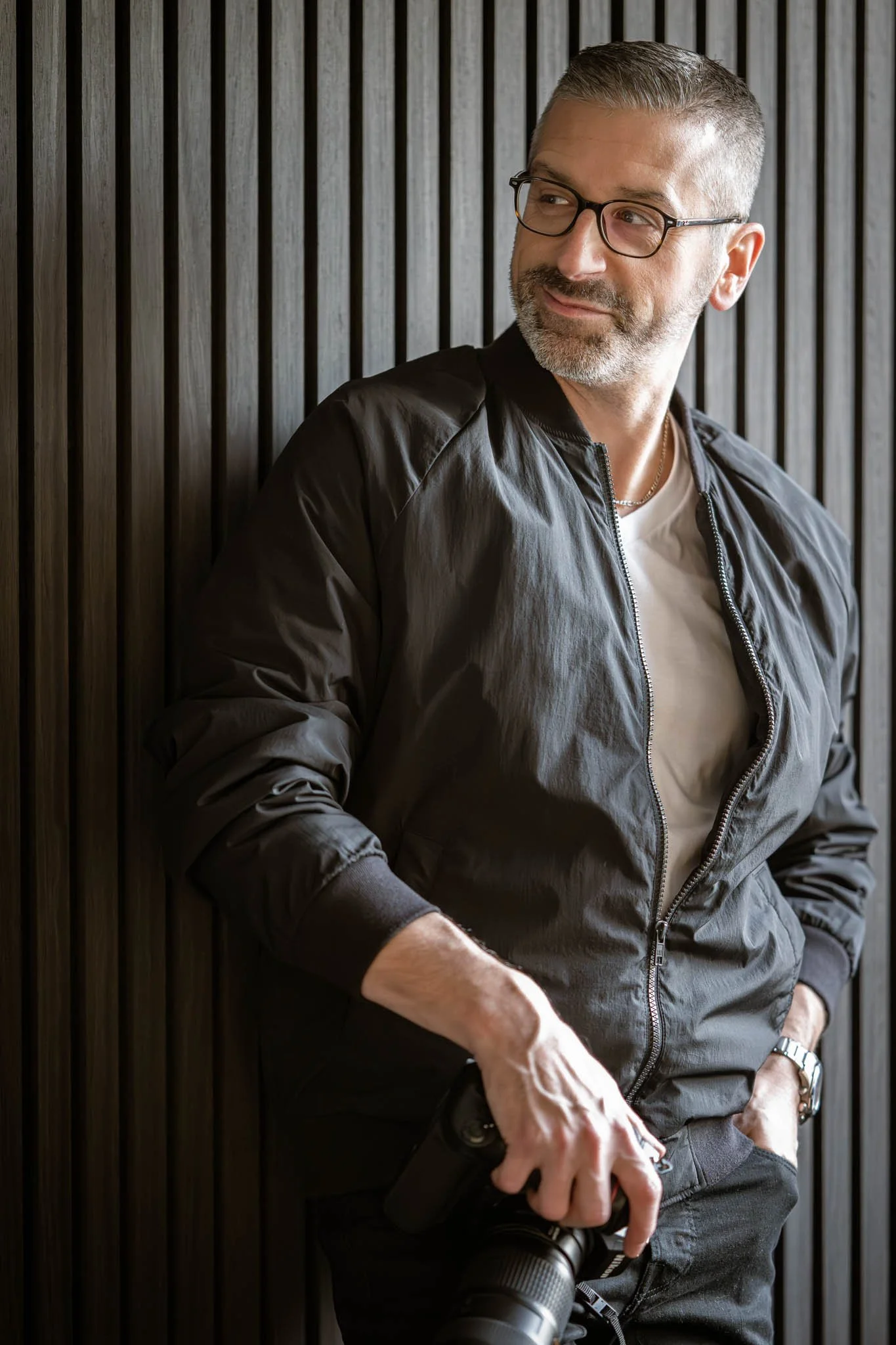 André Gosselin. Photographe de portrait. Photographe événement. Photo corporative.  Un homme portant des lunettes, une veste noire, une chemise blanche, tenant un appareil photo, posé contre un mur en bois vertical.