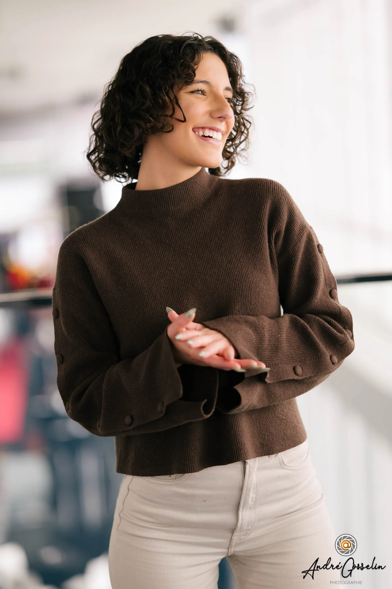 Portrait d'affaires corporatif. Une femme souriante portant un pull marron et un pantalon blanc, dans un intérieur lumineux.