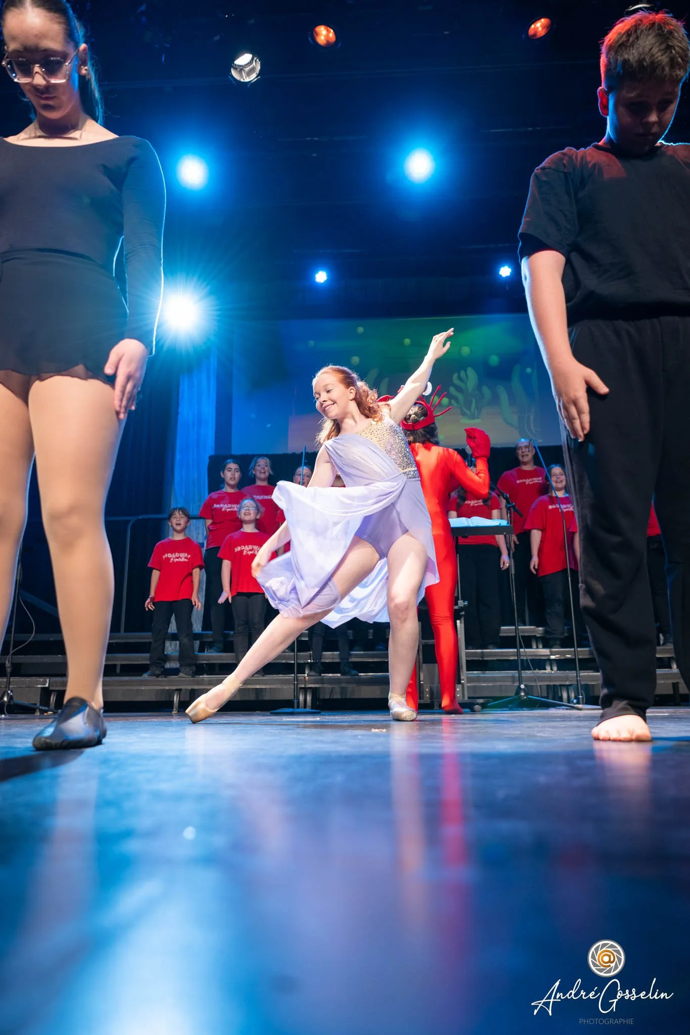Une scène de performance sur scène avec plusieurs danseurs et chanteurs. Au centre, une ballerine en tutu blanc effectue une pose de danse. En arrière-plan, un groupe de chanteurs en t-shirts rouges, un homme vêtu de noir et une femme en costume roug