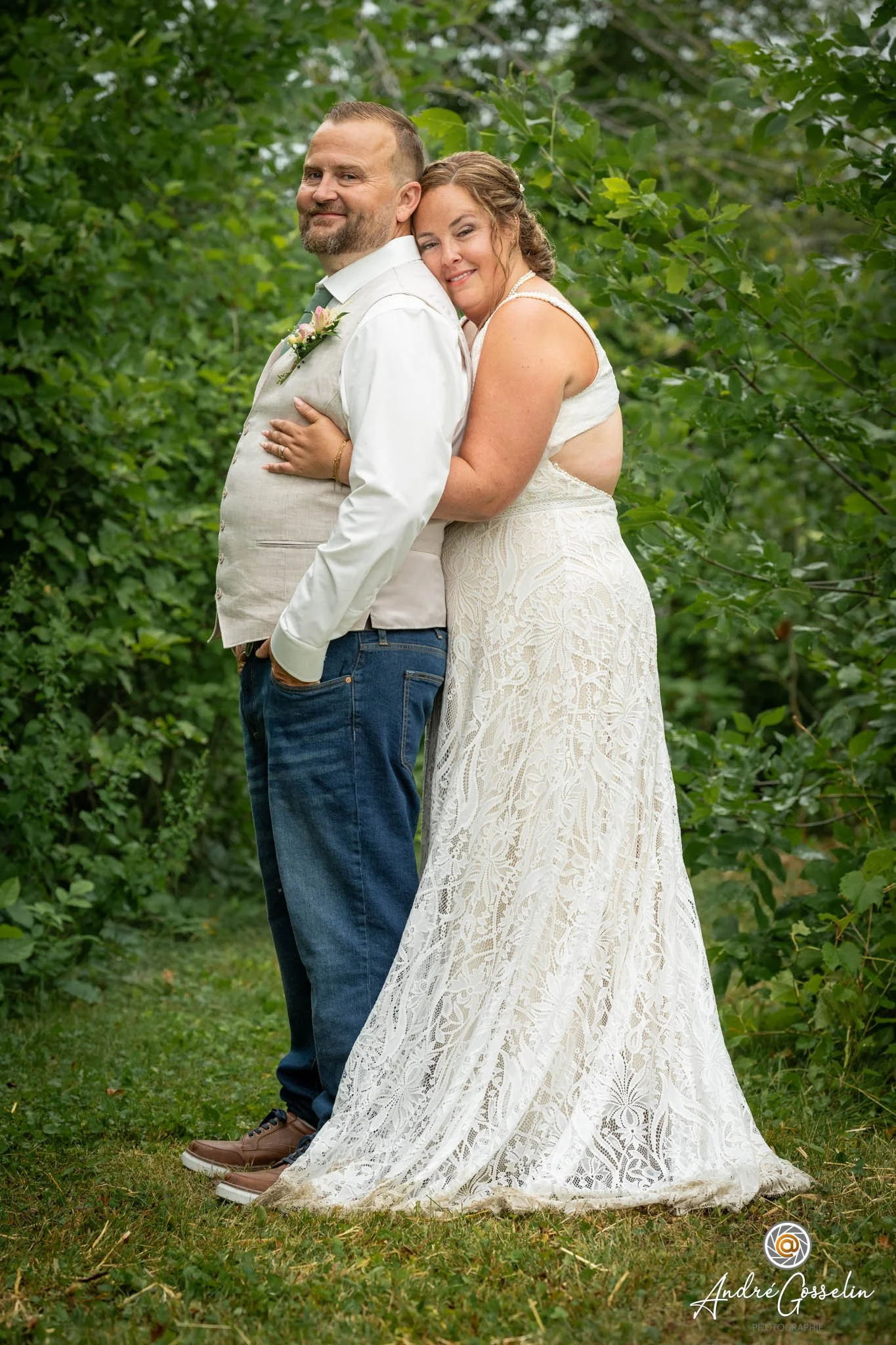 Un couple marié pose dans un cadre naturel avec des arbres et de l'herbe, la femme en robe de mariée et l'homme en costume décontracté, souriant et échangeant un regard affectueux.
