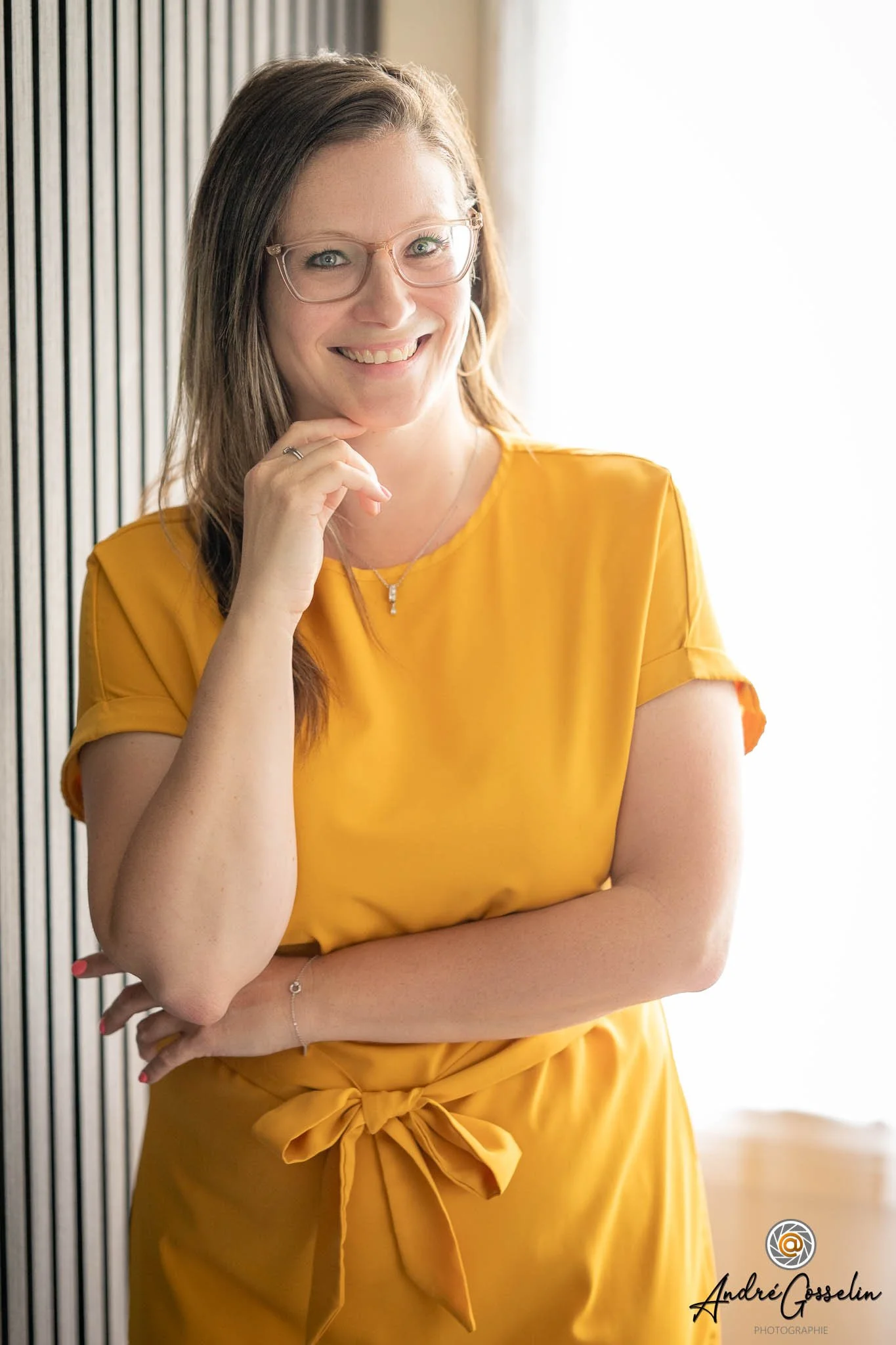 Une entrepreneure souriante portant des lunettes, vêtue d'une robe ou blouse jaune, posant devant un mur en bois et un rideau clair. Elle a les cheveux bruns, longs, et porte un collier et un bracelet fin. Photo corporative.