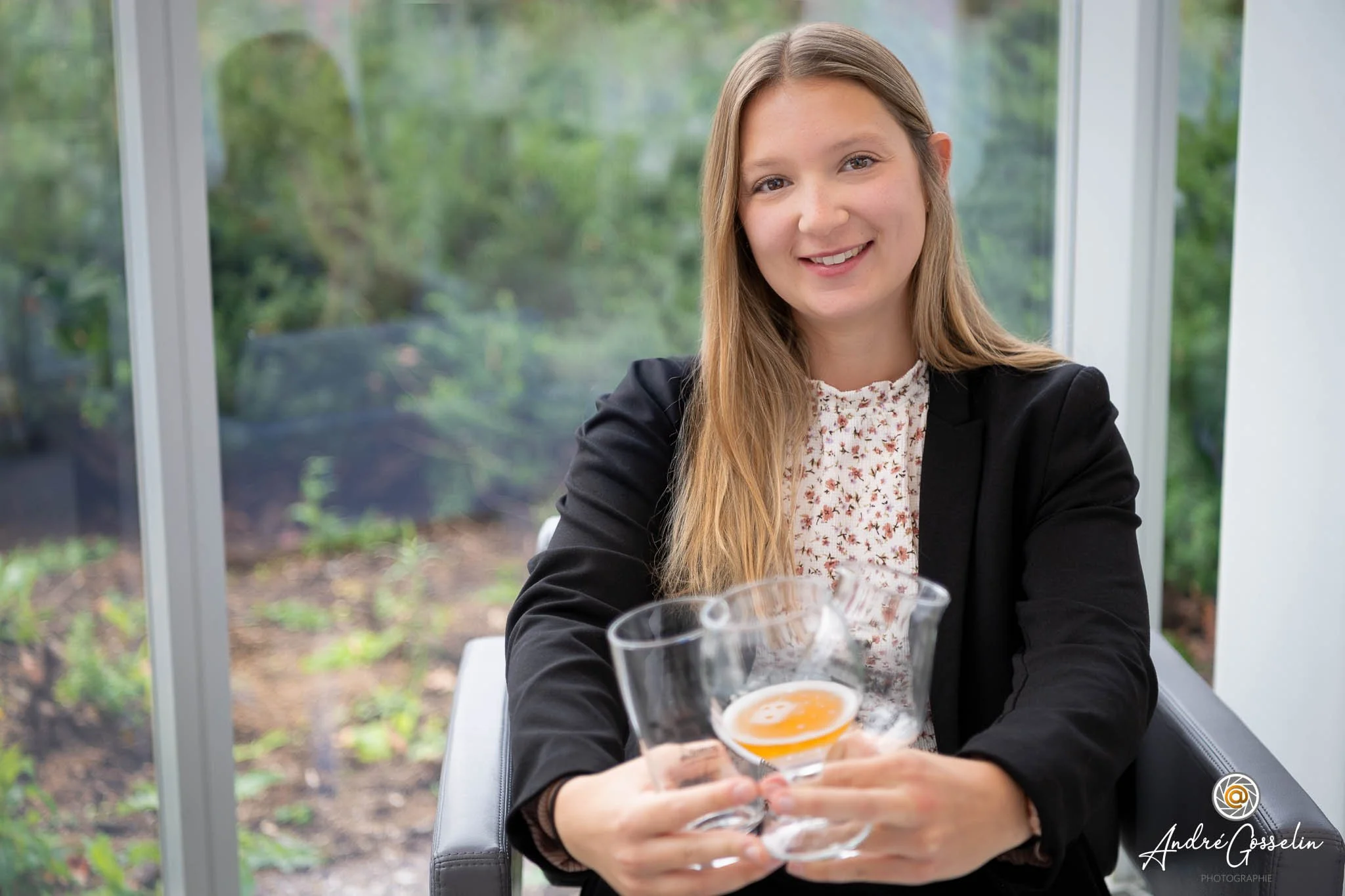 Une jeune femme souriante tenant un verre à cocktail avec une boisson orange, assise à une table près d'une fenêtre avec vue sur un jardin.