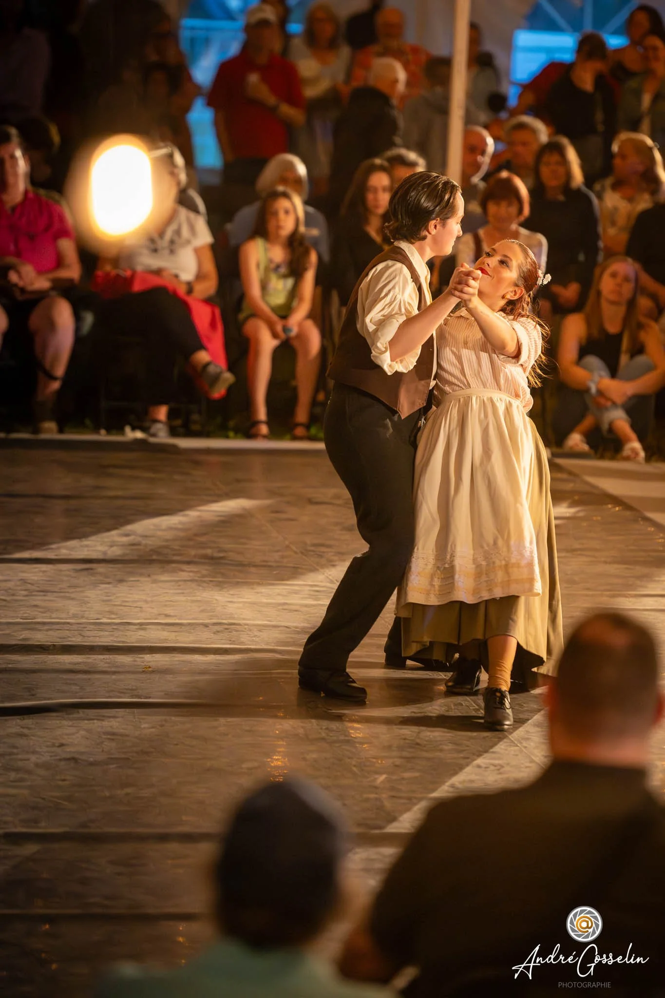 Deux jeunes danseurs dansent lors d'une représentation théâtrale ou une scène de théâtre, avec un public assis en arrière-plan, dans une ambiance chaleureuse et intimiste.