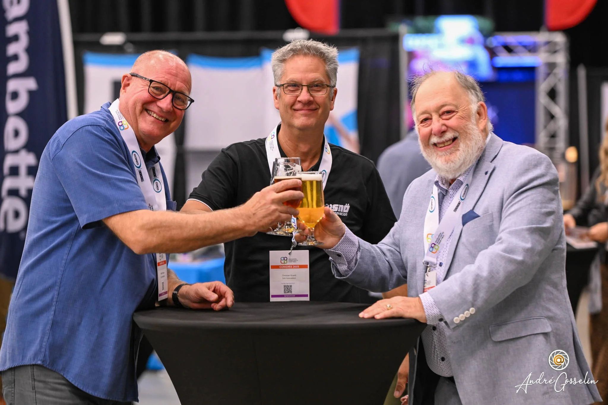 Trois hommes souriants, en costume et tenues décontractées, trinquent avec des verres de bière lors d'un événement professionnel, un à une table haute.