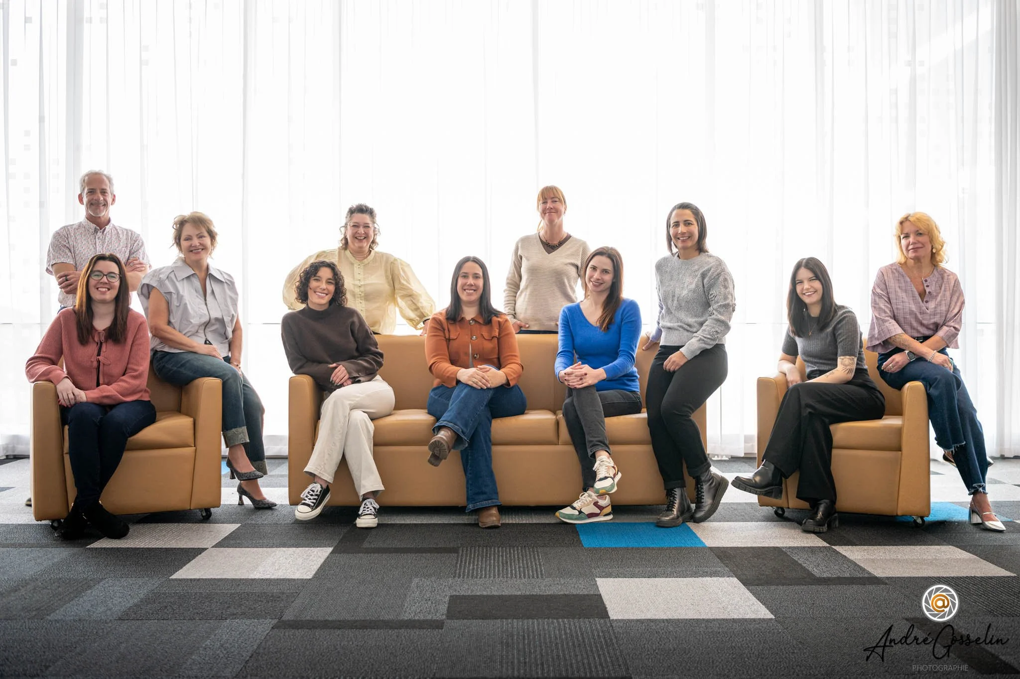 Photo corporative de groupe style lifestyle. Dix personnes rassemblées dans un espace lumineux avec de grandes fenêtres derrière elles, assises et debout, souriant à l'appareil.