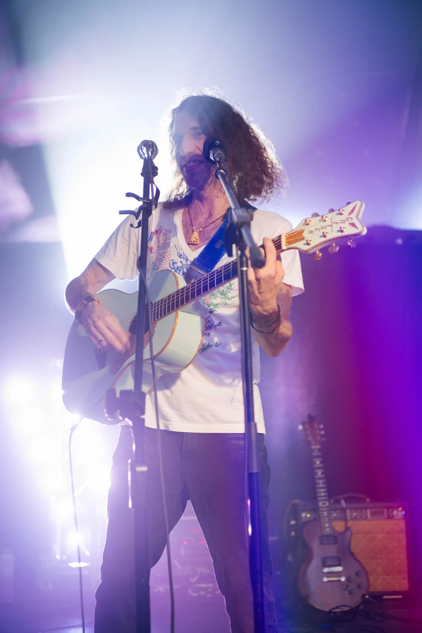 Un homme avec des cheveux bouclés et une barbe tenant une guitare acoustique, en train de chanter ou jouer lors d'une performance sur scène, entouré d'effets de lumière violette et bleue. Portrait de spectacle.