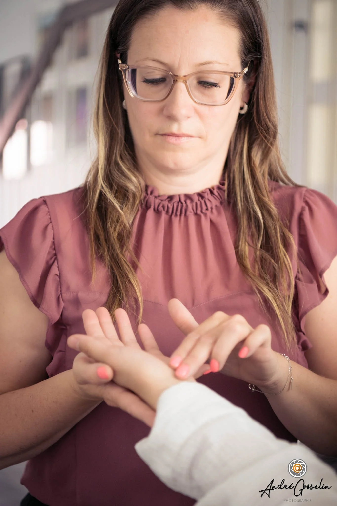 Photo corporative. Une femme porte des lunettes et une blouse rose, ses mains sont croisées devant elle, un enfant tend la main vers elle. Image claire, ambiance chaleureuse.