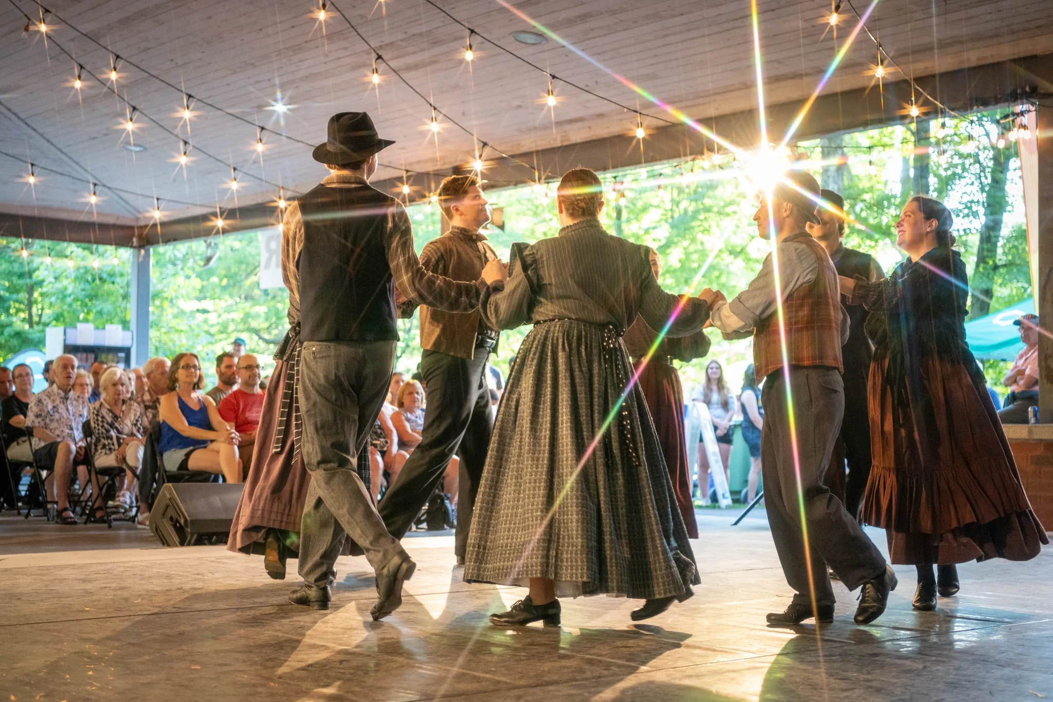 Groupe de personnes dansant en costumes traditionnels lors d'une fête en plein air, avec un public assis et un éclairage festif en soirée. Photo de spectacle.