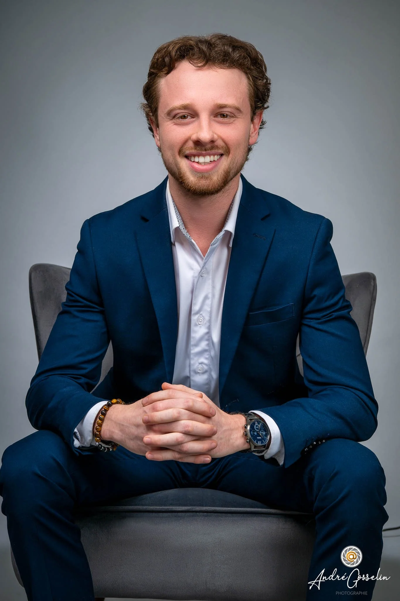 Un homme souriant en costume bleu, assis sur une chaise, avec une montre et un bracelet, face à un fond neutre. Portrait corporatif. Photo professionnelle.