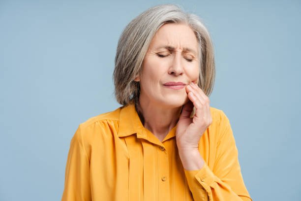An elderly woman with gray hair in a yellow blouse holds her jaw with a pained expression, indicating tooth or jaw pain, against a light blue background.