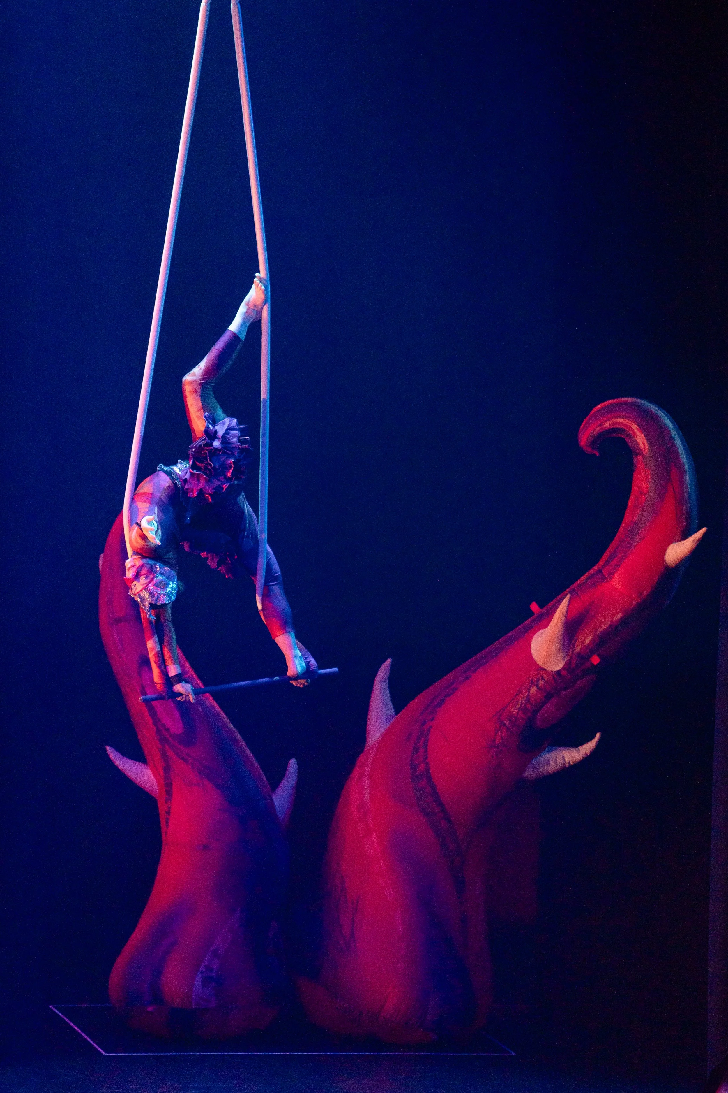 Circus performer dressed in colorful costume balancing on a pole over a large fake dinosaur with red skin and white horns.