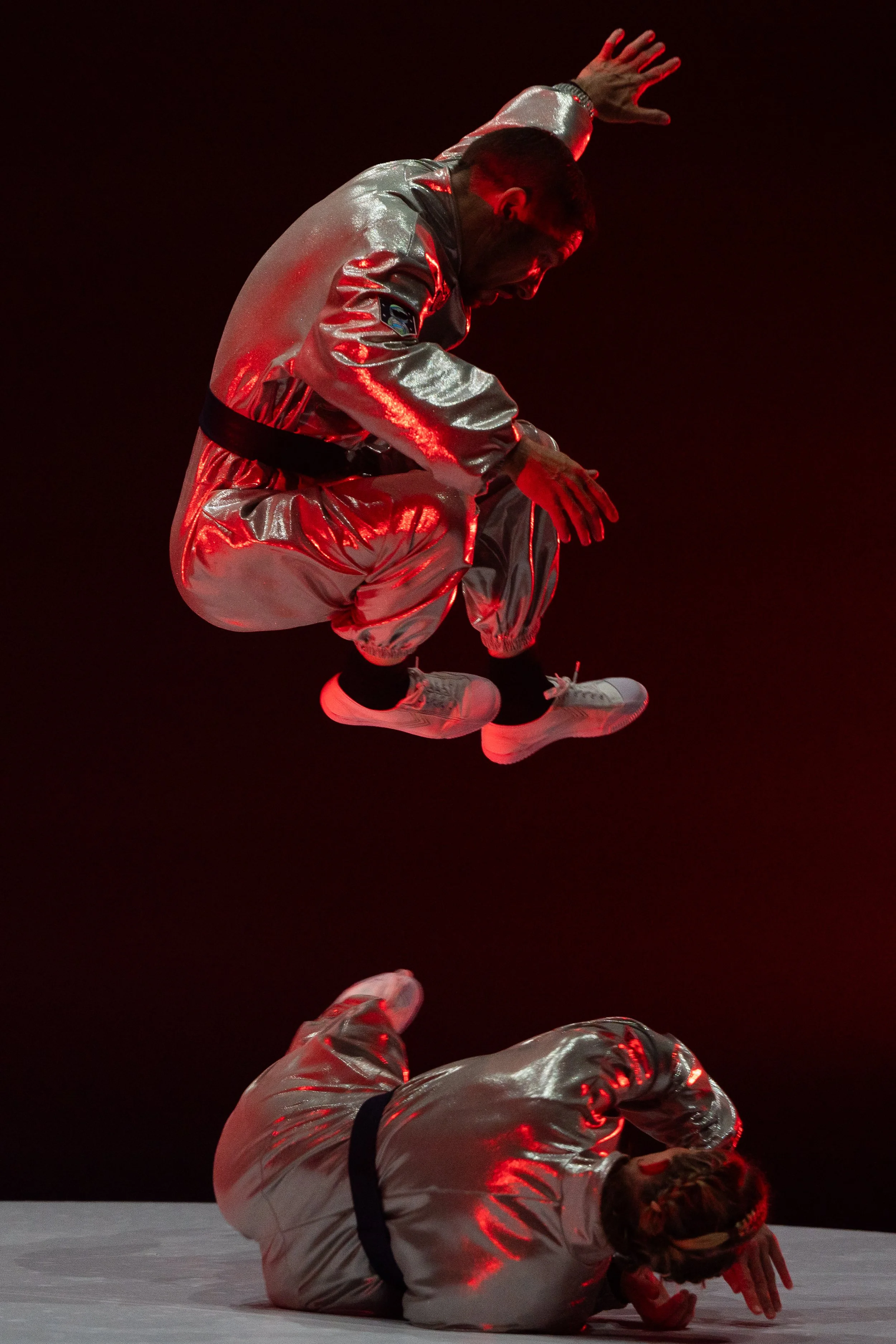 Two performers in shiny silver outfits are captured mid-performance against a dark background. One is airborne, crouching with arms raised and head bent forward, while the other is lying on the ground in a similar outfit, with arms covering the head.
