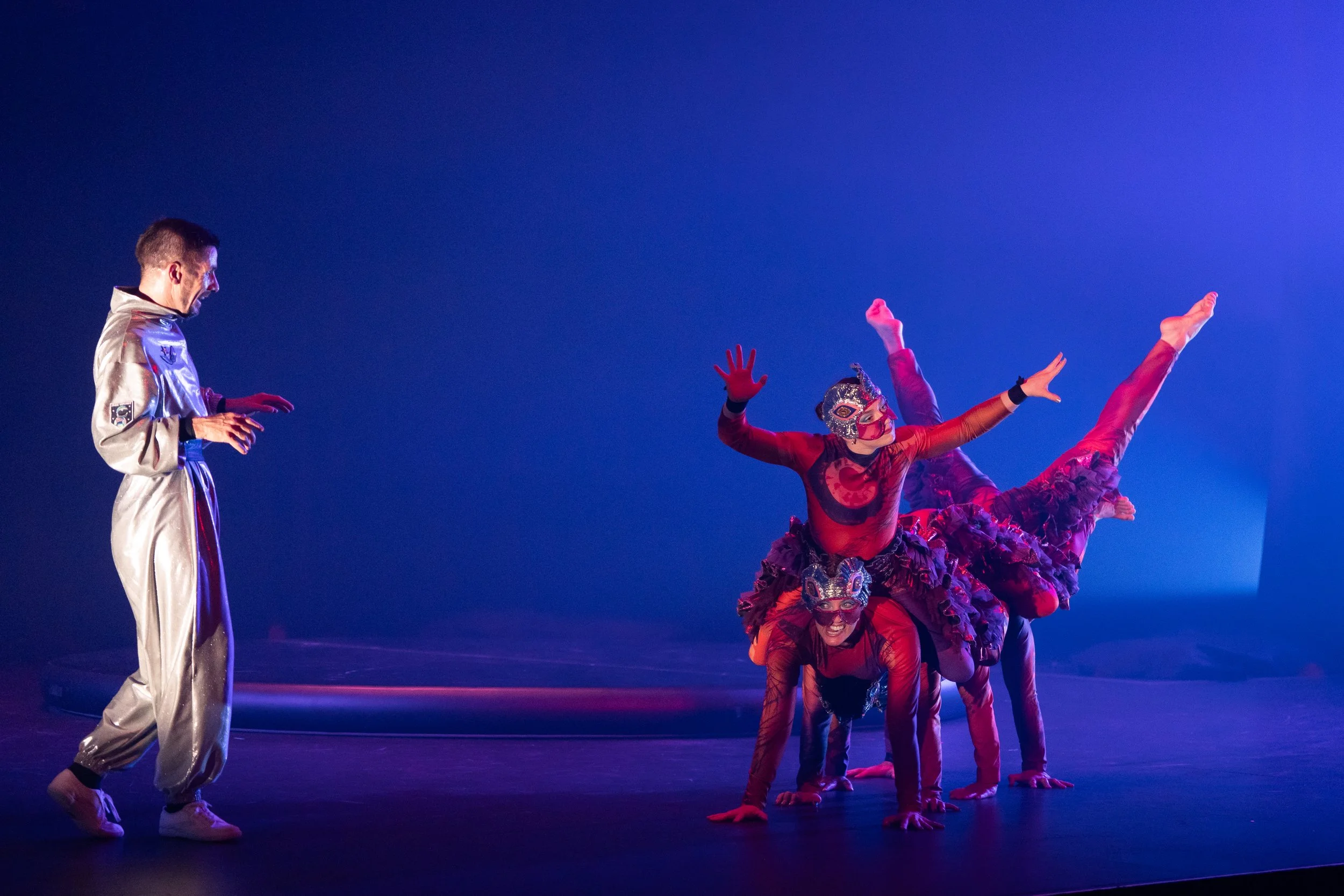 Circus performers in colorful costumes, one man in silver standing to the left, three women in red and purple masks and costumes performing acrobatic poses on stage, illuminated with blue and purple lighting.