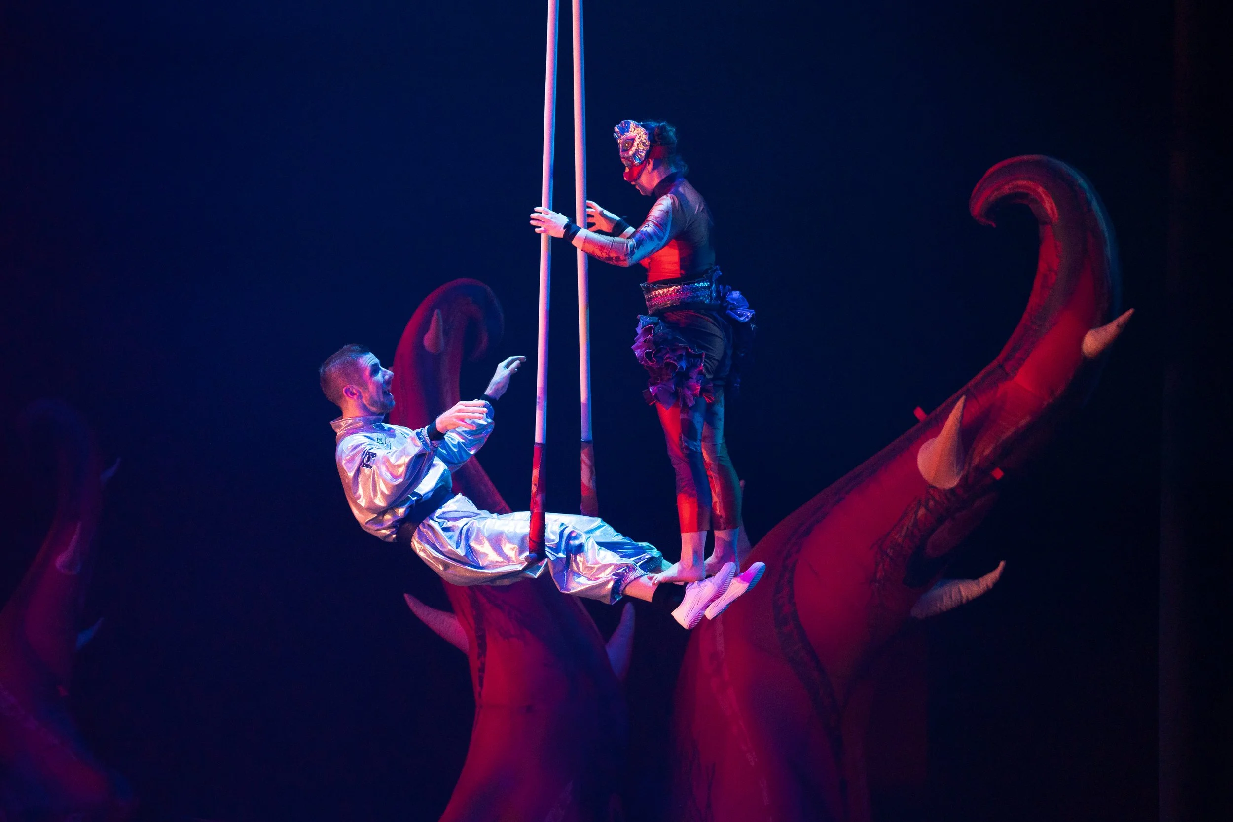 Two performers in colorful costumes on a stage, one sitting on a red dinosaur prop, the other standing on the dinosaur's back, holding onto poles, with dark background and dramatic lighting.