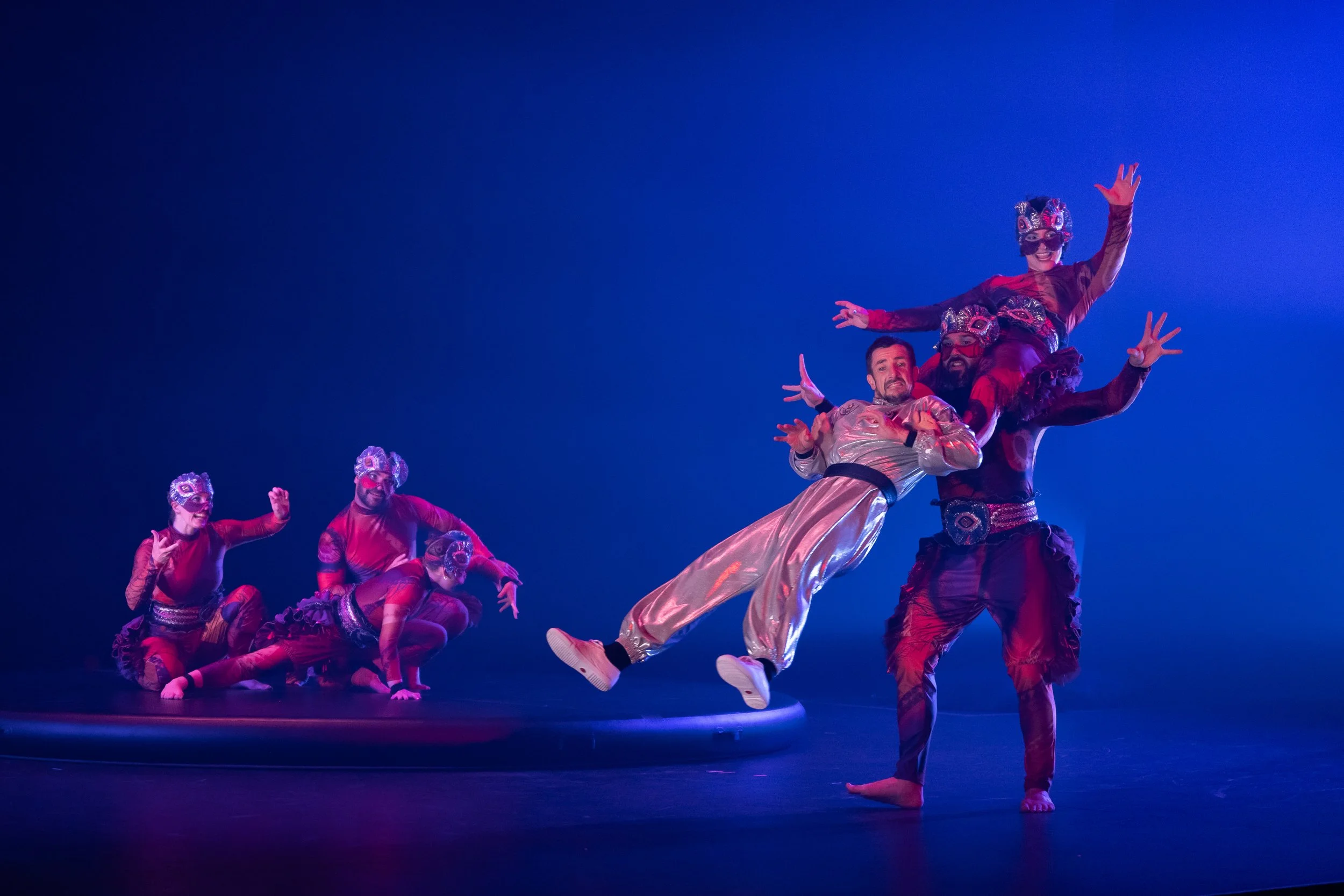 Performers in colorful costumes and masks dancing on stage during a theatrical performance with a blue background.