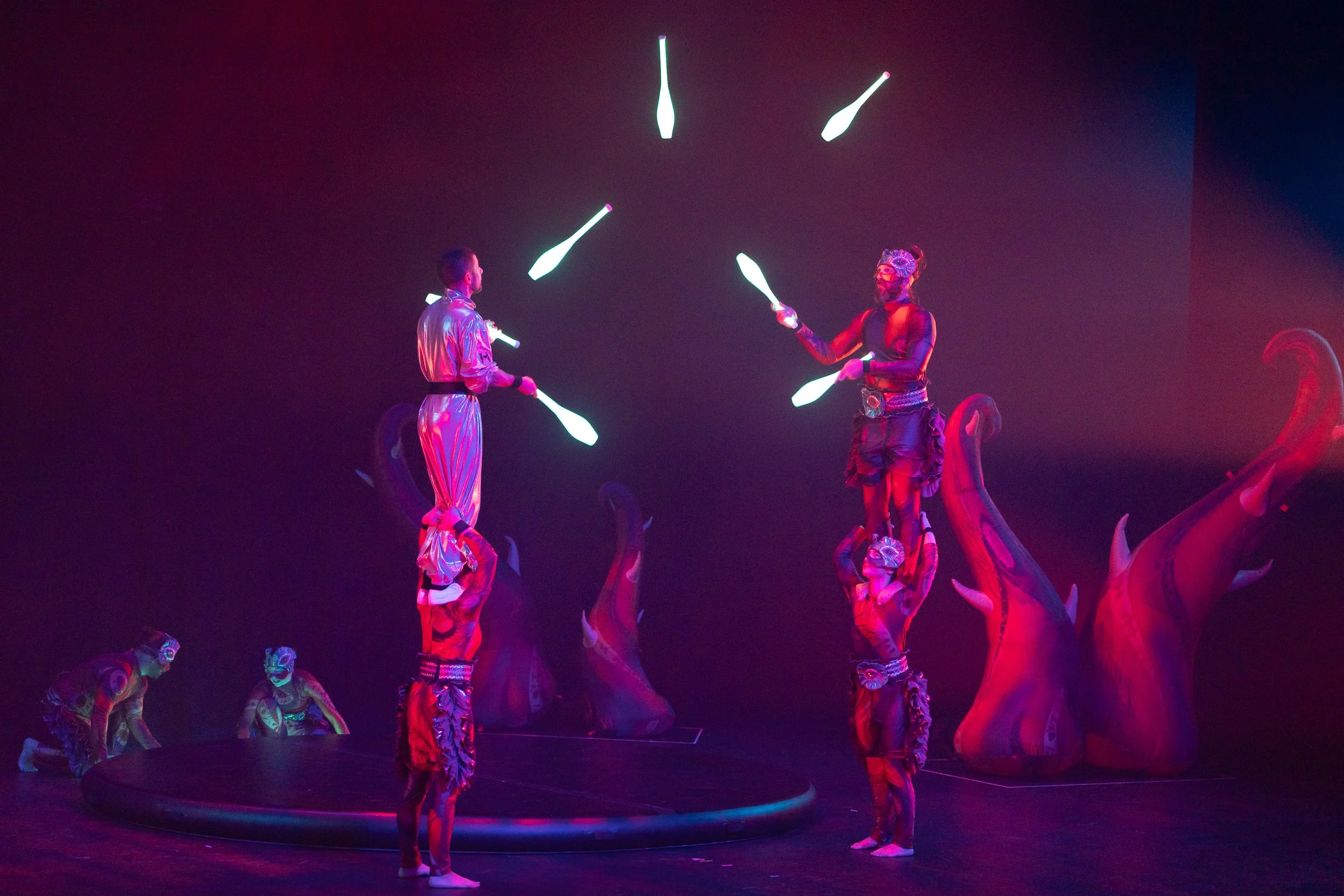 Performers juggling glowing clubs on a stage with large red dinosaur sculptures and purple lighting.