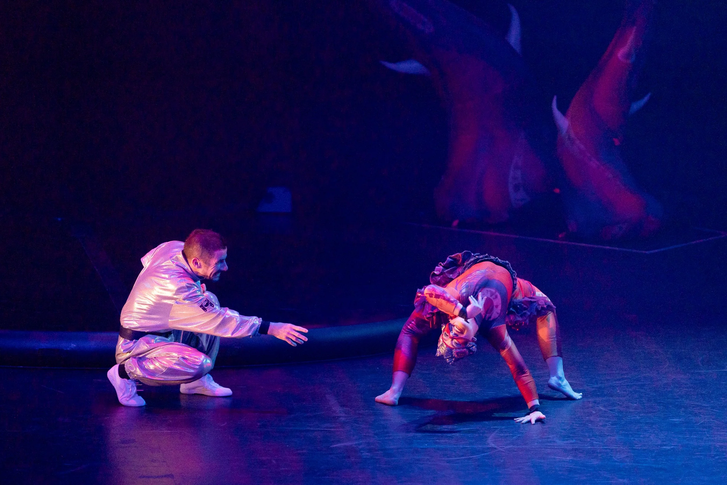 A man in a shiny silver jacket and pants kneels on stage, reaching out towards a woman in a colorful, patterned bodysuit who is bent over with one hand on the ground and the other hand pointing upward, both performing a dance routine with a dark back