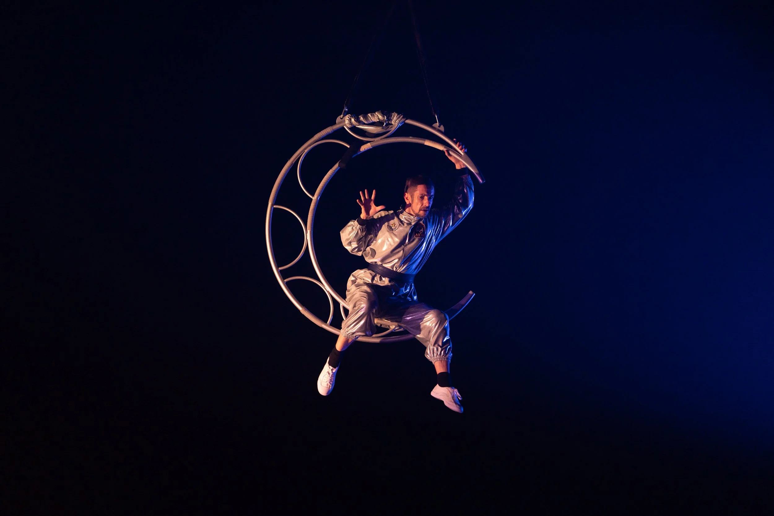 A performer in a shiny silver costume is sitting in a crescent moon-shaped metal apparatus suspended in the air against a dark background, possibly during a nighttime circus or aerial act.