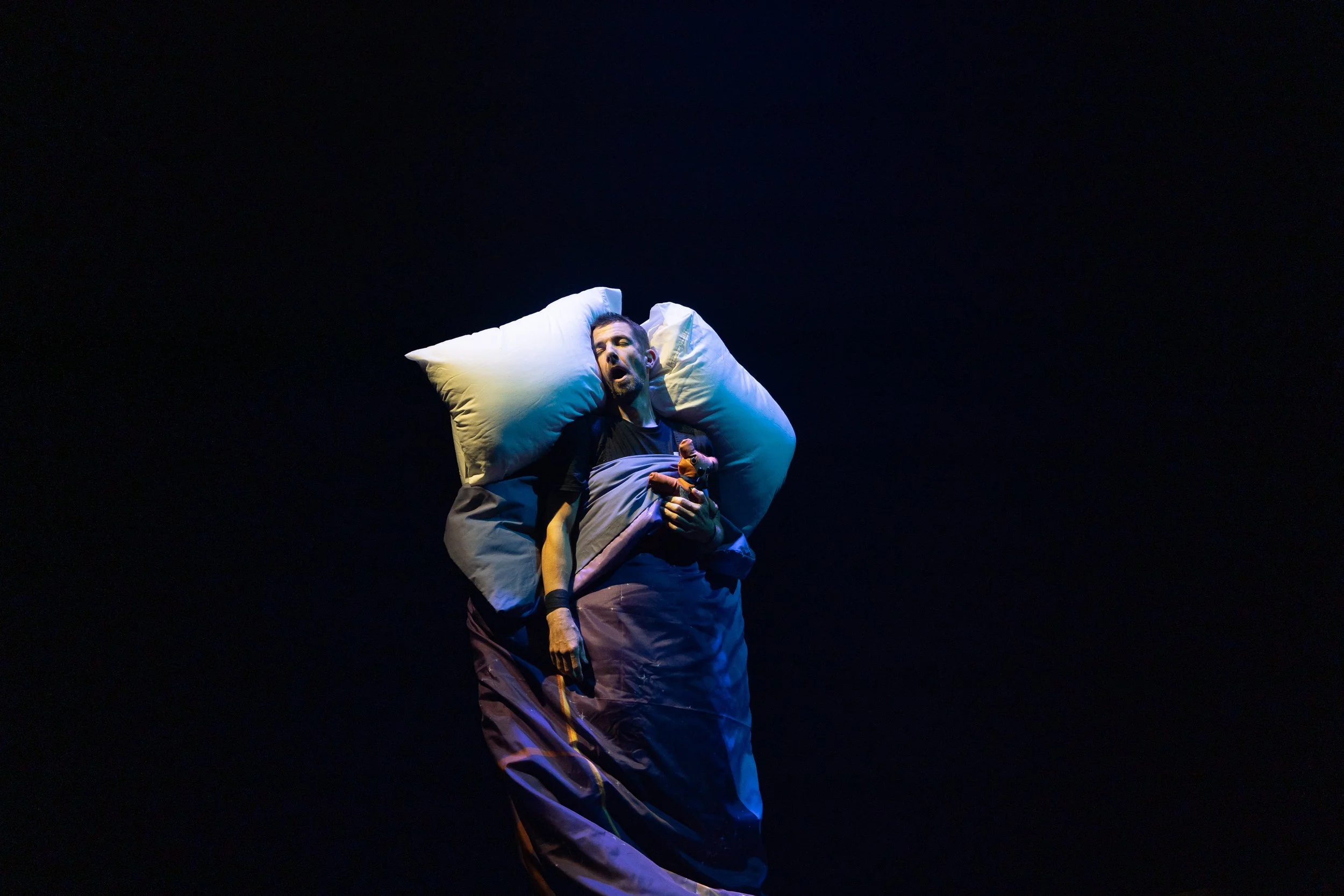 A man lying on a bed with a pillow, singing or speaking with a microphone, set against a dark background.