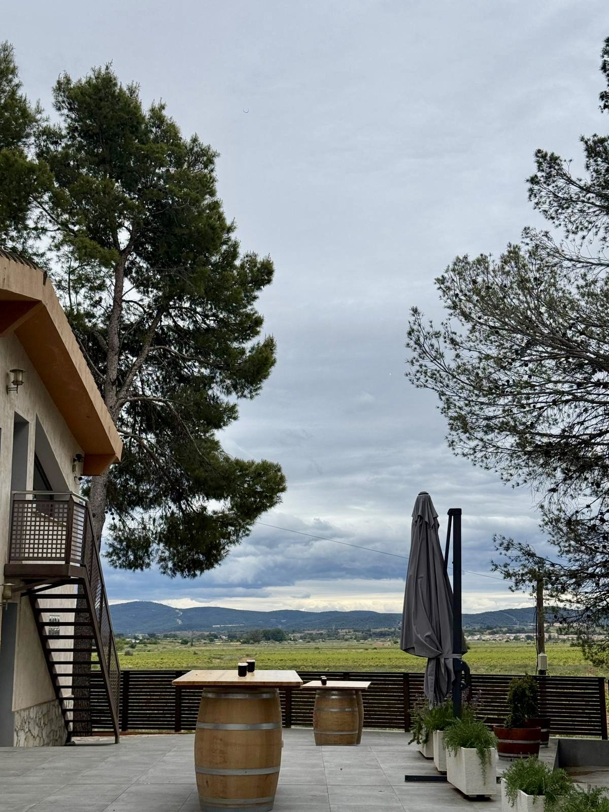 Outdoor patio area with two wooden barrel tables, a large umbrella, and potted plants, overlooking a scenic landscape with mountains and an overcast sky.