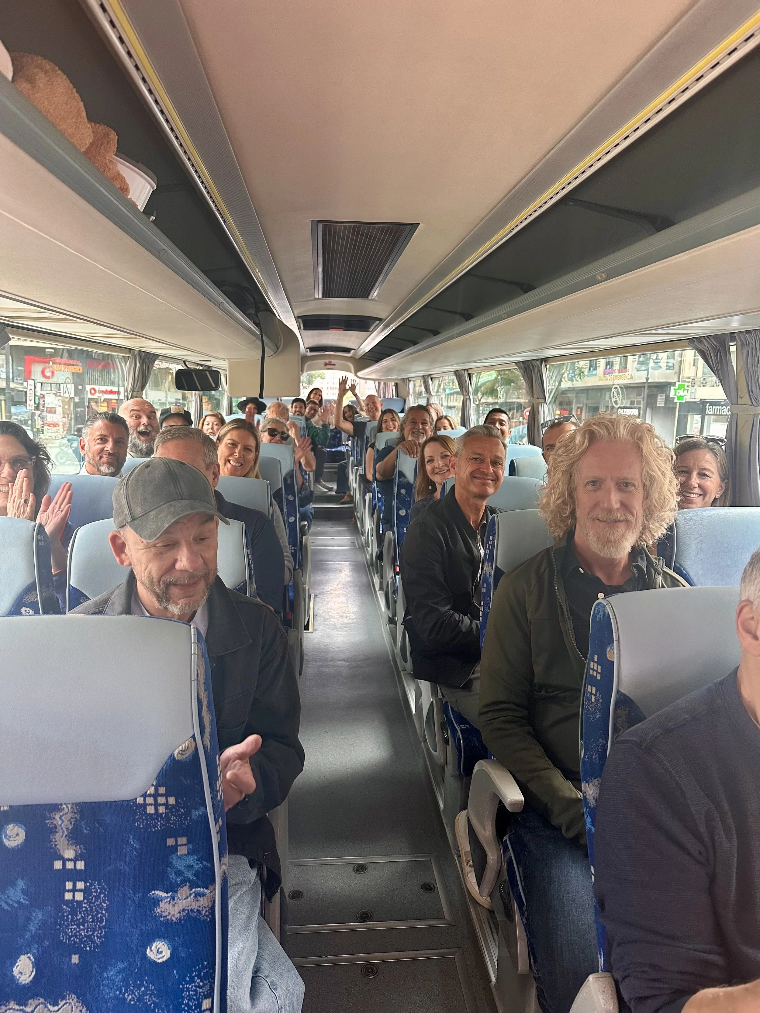 People sitting in a bus, with some smiling and waving at the camera, showing a cheerful atmosphere.