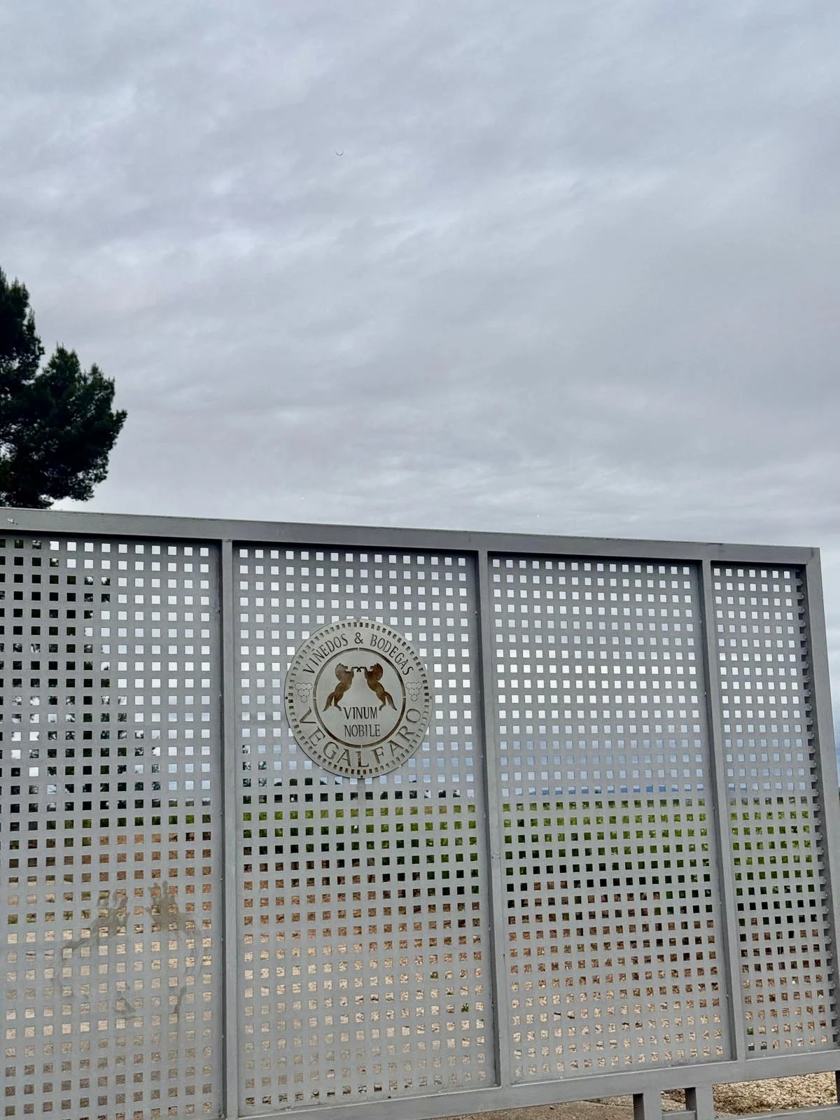 A metal fence with a circular logo in the center, featuring two dogs and the words 'Vinedos & Bodega,' 'Vinum Nobile,' and 'Vegal Fardo' on a cloudy day.
