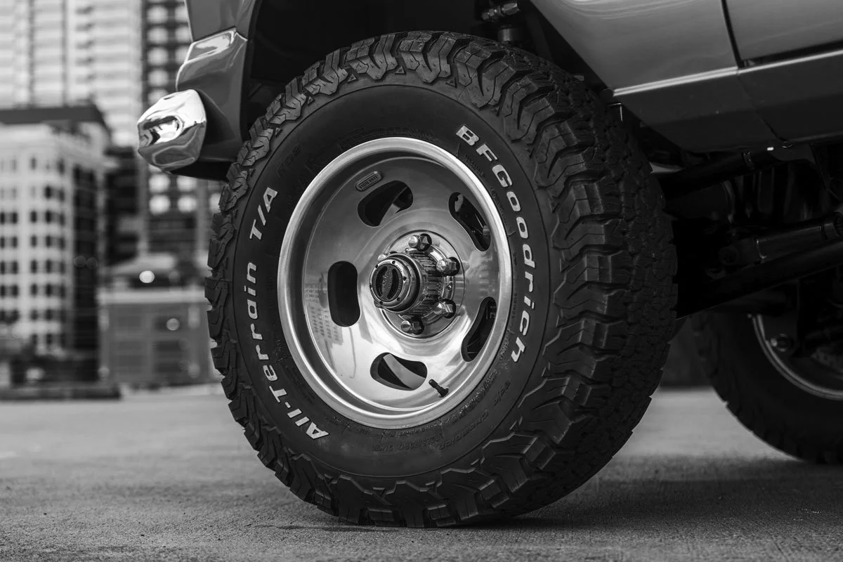 Close-up of a rugged off-road tire on a vehicle, with city buildings in the background, in black and white.