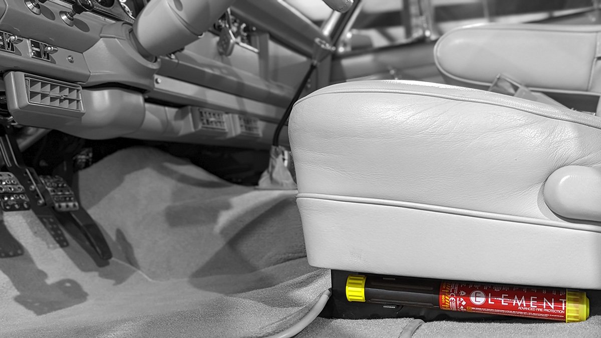 Inside a vehicle, view of the driver’s seat, pedals, and part of the dashboard with a fire extinguisher labeled "ELEMENT" mounted underneath the seat.