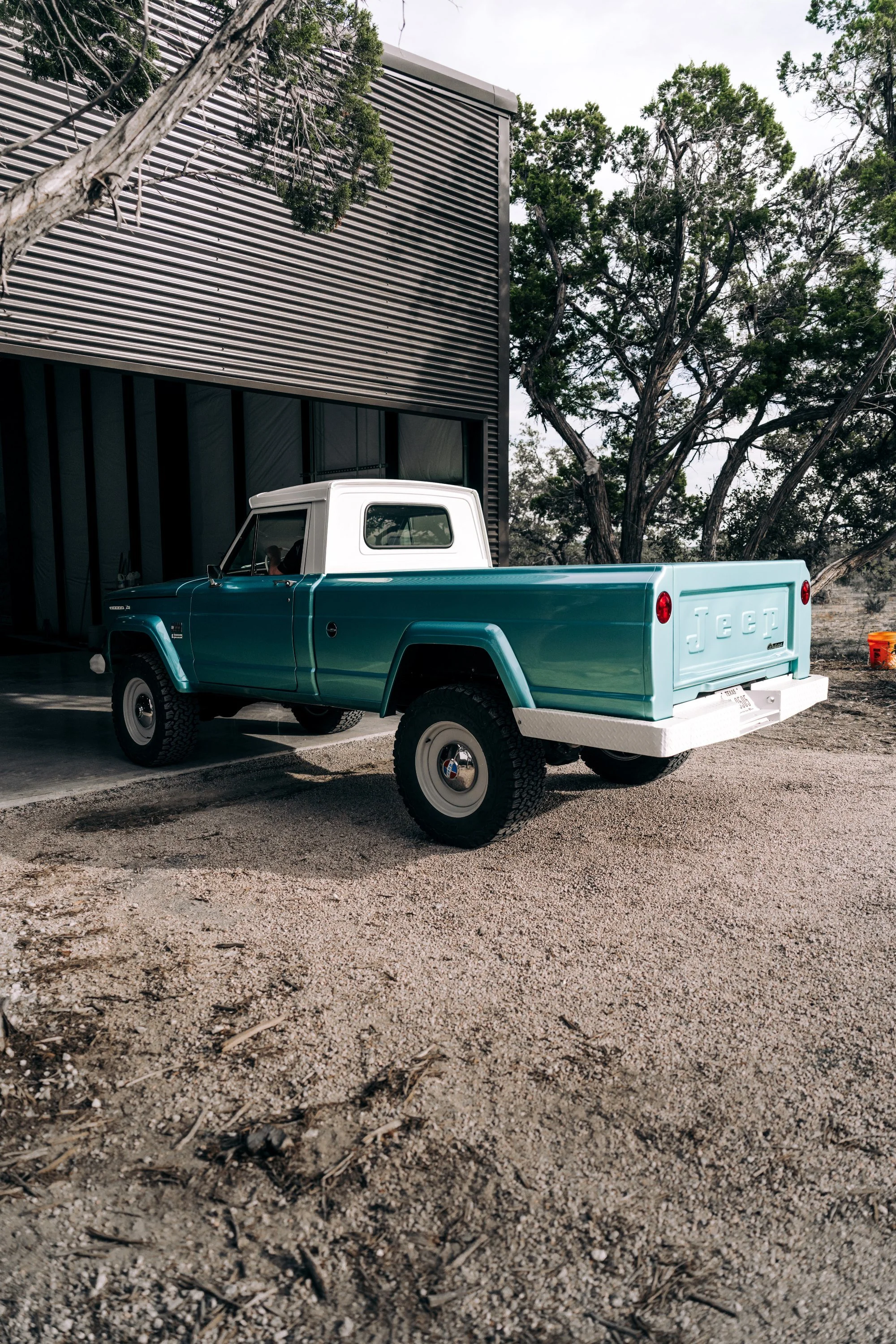 A vintage turquoise pickup truck with a white camper shell parked outside a modern black building with large windows and surrounded by trees.