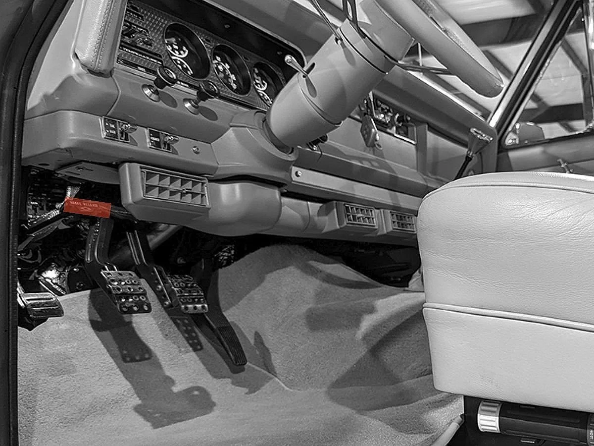 Interior of a classic car showing the dashboard, steering wheel, gas and brake pedals, and part of a white leather seat.