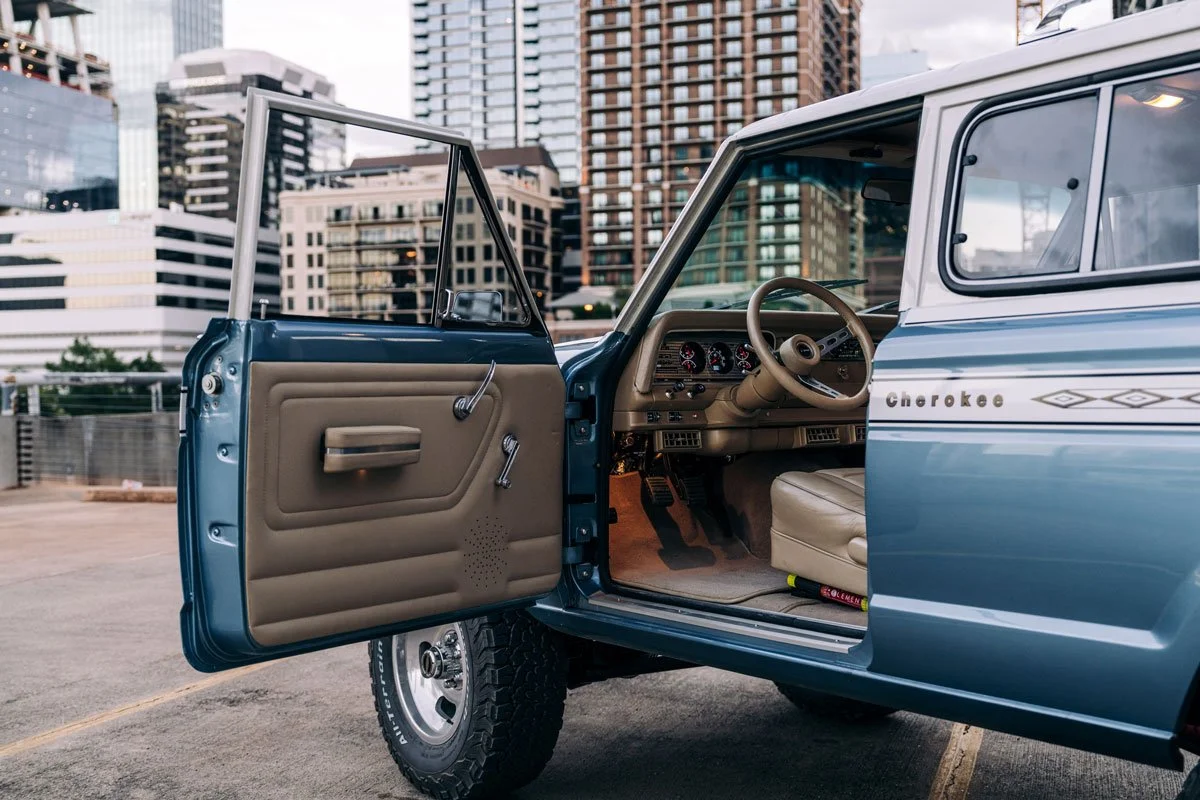 Inside view of a vintage blue Jeep Cherokee with the driver side door open, showing the beige interior, steering wheel, dashboard, and city buildings in the background.