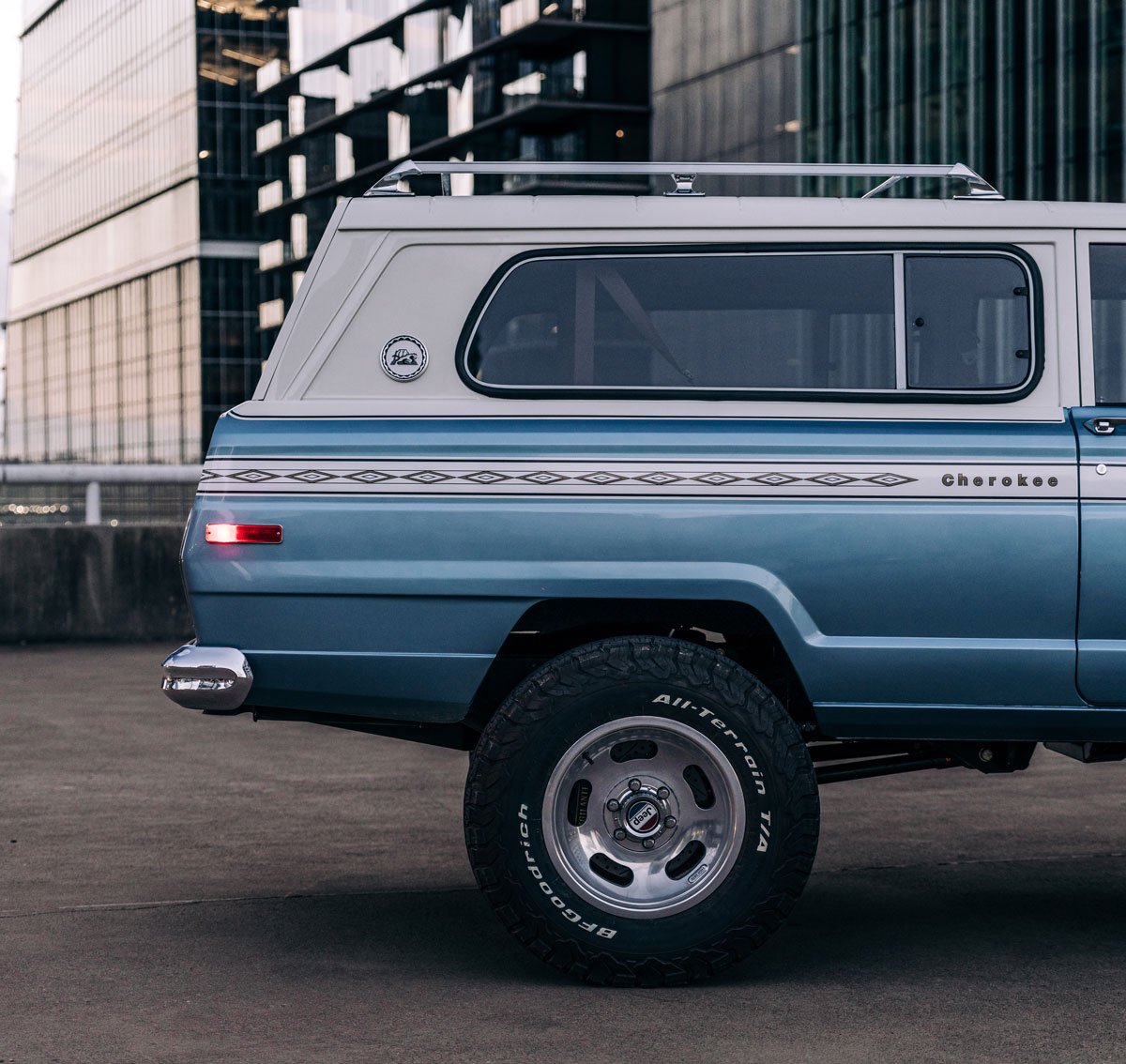Side view of a vintage Cherokee SUV with a two-tone blue and white color scheme parked near modern glass buildings.