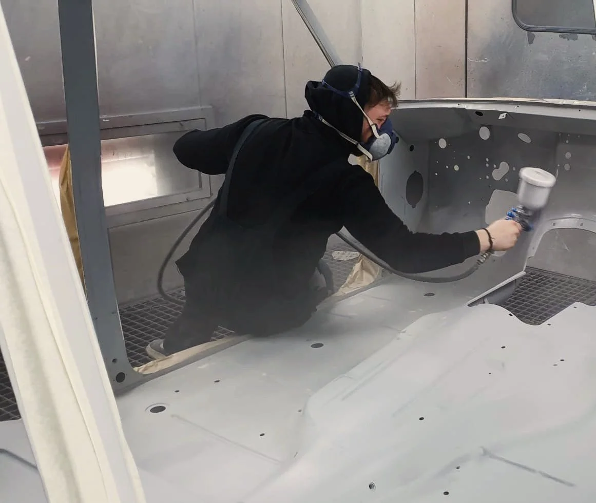 Person wearing a mask and protective gear spray painting the interior of a metal aircraft fuselage or similar structure in a spray booth.