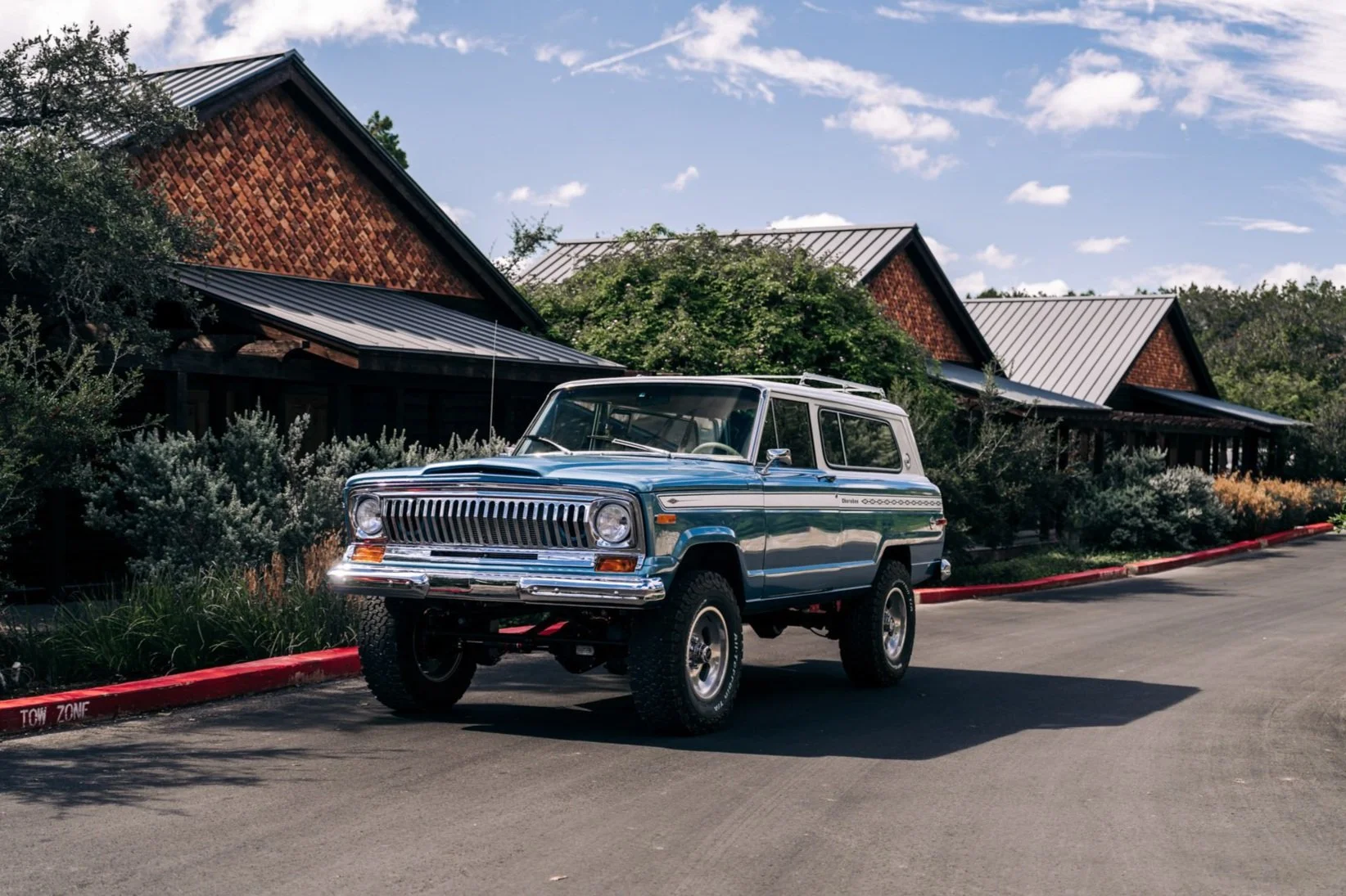 A vintage blue Jeep parked on a paved driveway next to a landscaped area with bushes and greenery. In the background, there are buildings with sloped roofs and a partly cloudy blue sky.