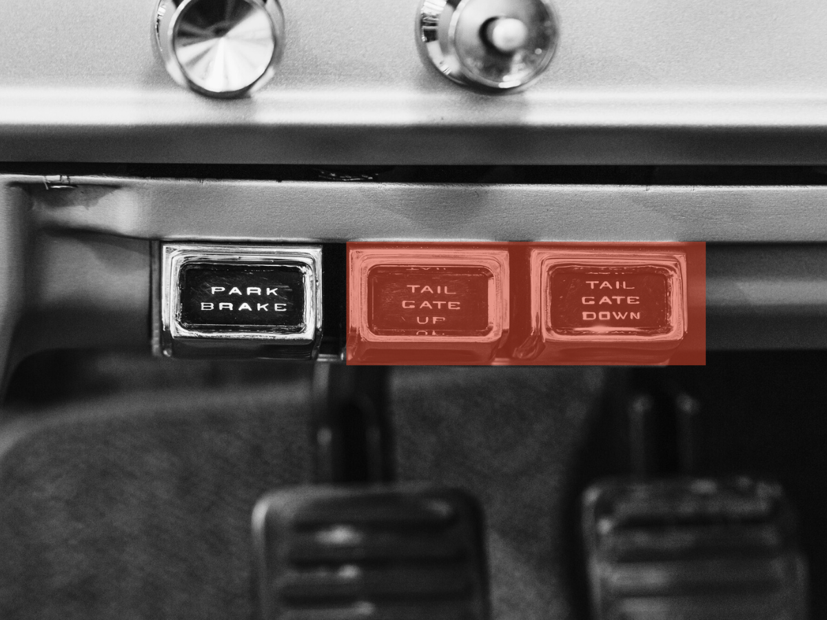 Close-up of a control panel with a black button labeled 'Park Brake' and two red buttons labeled 'Tail Gate Up' and 'Tail Gate Down' in an industrial or transportation vehicle.